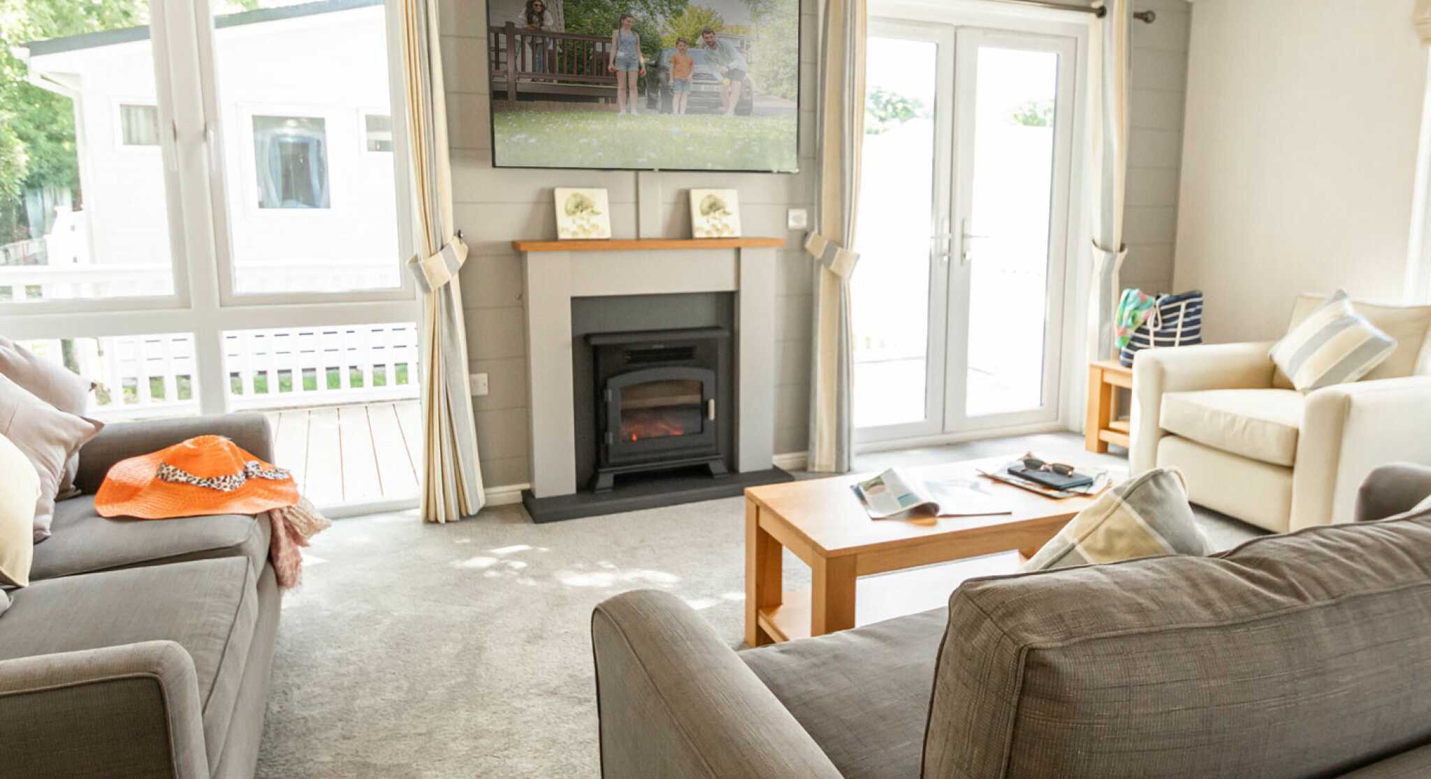 A cozy living room featuring two sofas, a coffee table, and a television mounted on the wall. Light streams in through large windows, with a view of a deck outside. Soft, neutral colors and decorative pillows create a warm atmosphere.