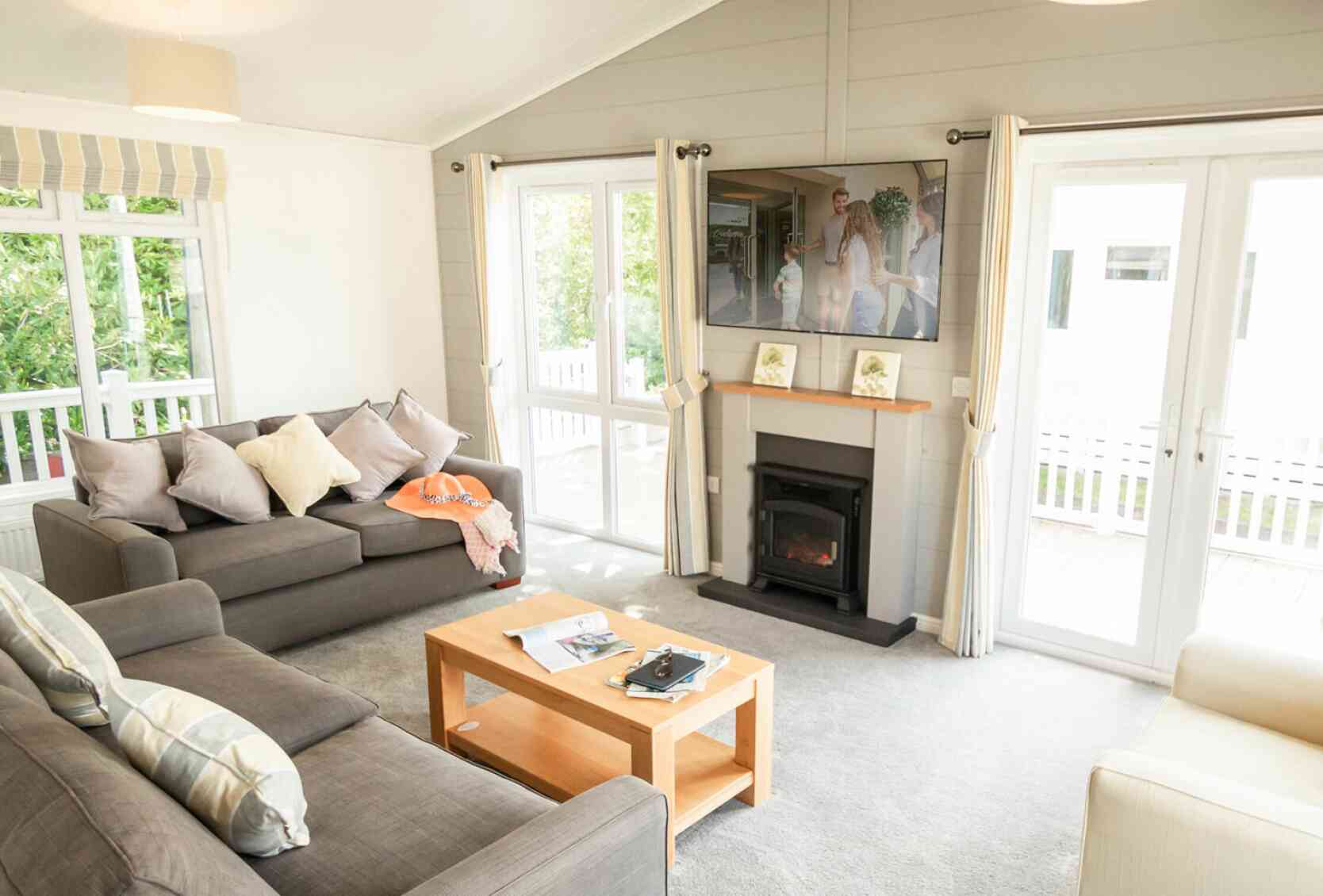 A bright living room features two gray sofas with decorative pillows, a wooden coffee table with magazines, and a flat-screen TV mounted on the wall. Large windows let in natural light, and sliding glass doors lead to an outdoor area.