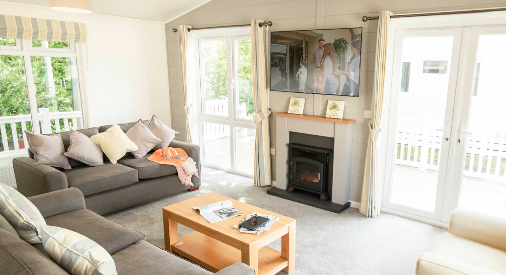 A bright living room features two gray sofas with decorative pillows, a wooden coffee table with magazines, and a flat-screen TV mounted on the wall. Large windows let in natural light, and sliding glass doors lead to an outdoor area.