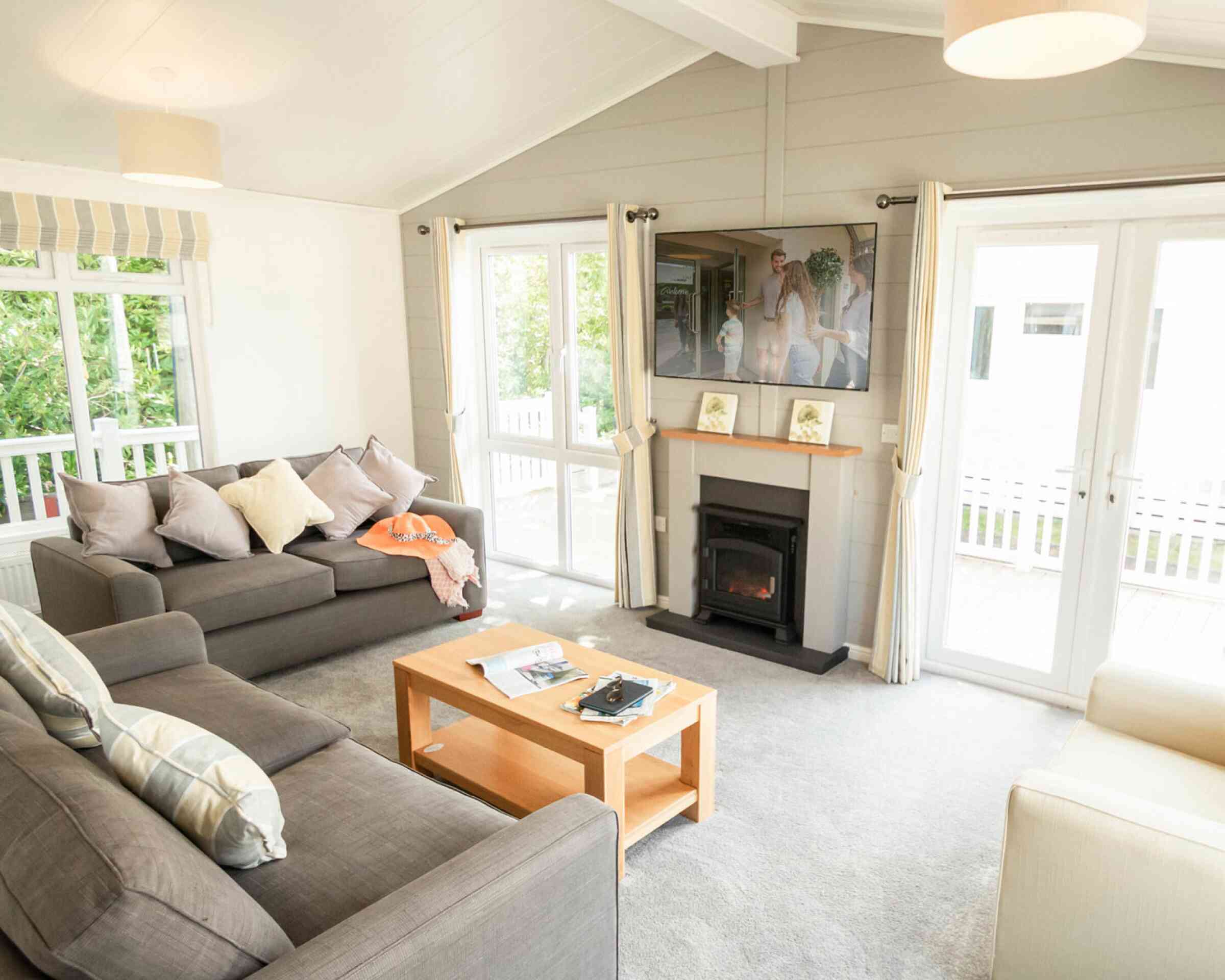 A bright living room features two gray sofas with decorative pillows, a wooden coffee table with magazines, and a flat-screen TV mounted on the wall. Large windows let in natural light, and sliding glass doors lead to an outdoor area.