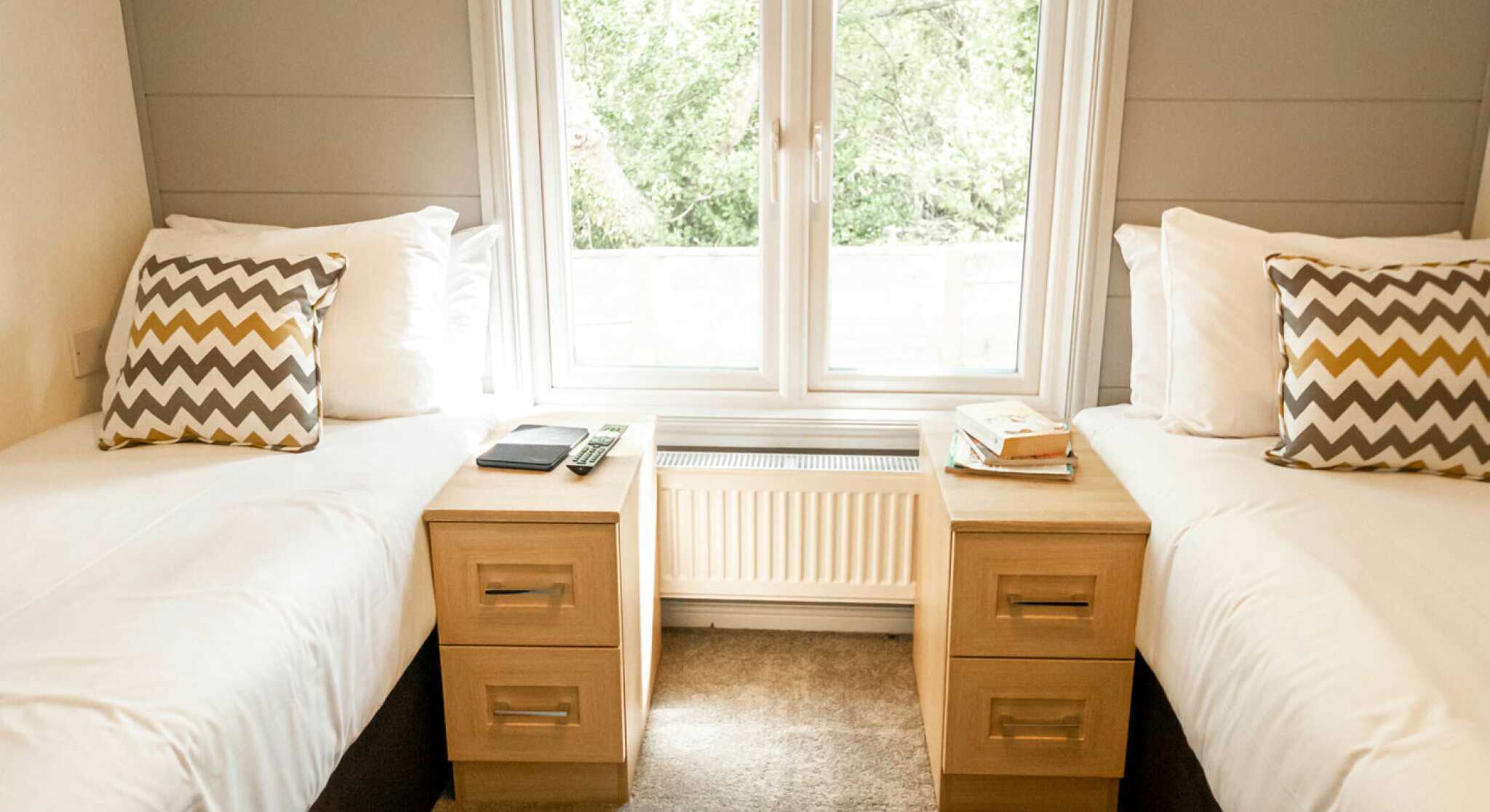 Two twin beds with white bedding and decorative chevron-patterned pillows, positioned next to each other. Each bed has a wooden nightstand with a book and a clock on top, set against a large window that offers a view of greenery outside. The floor is carpeted in a neutral color.