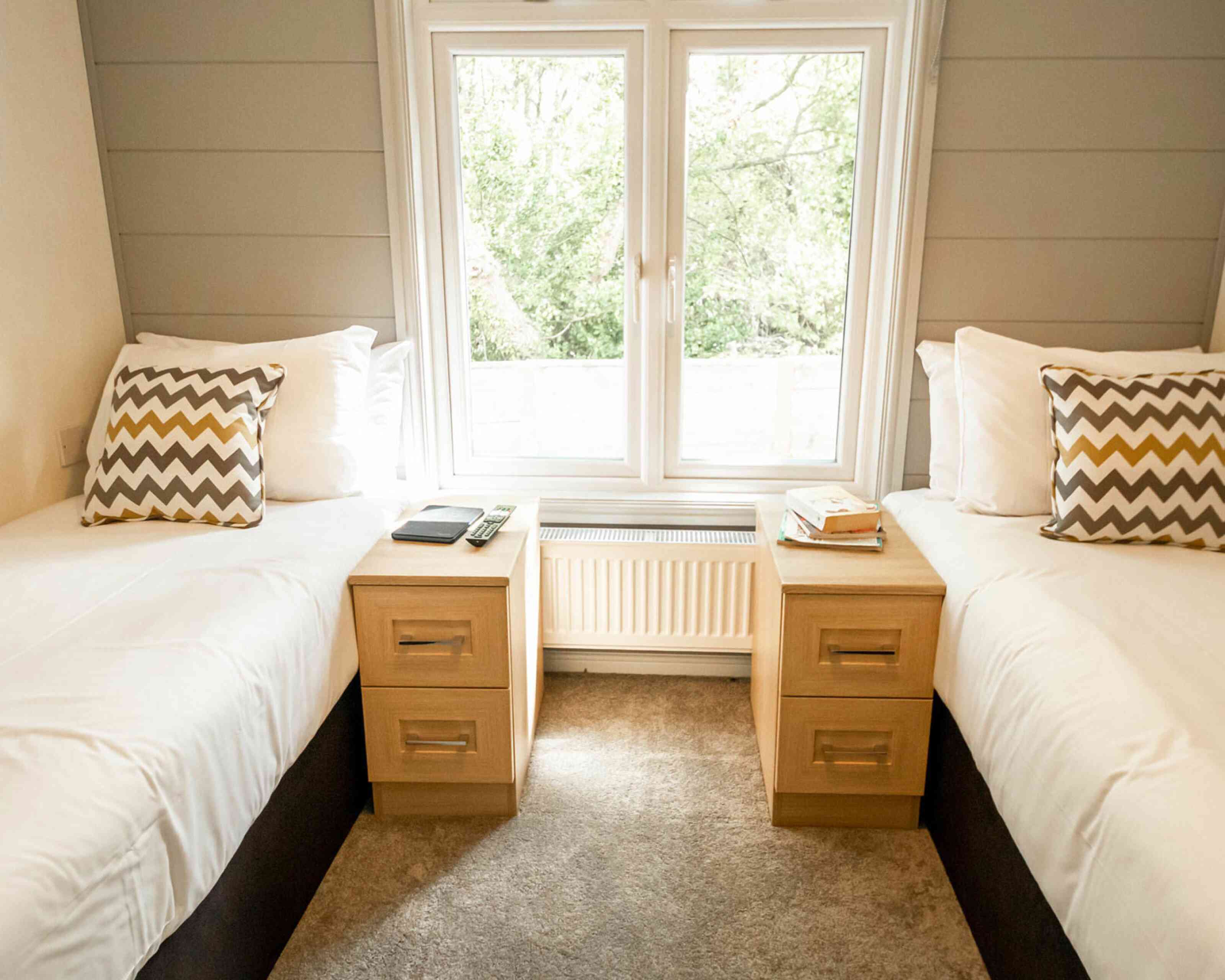 Two twin beds with white bedding and decorative chevron-patterned pillows, positioned next to each other. Each bed has a wooden nightstand with a book and a clock on top, set against a large window that offers a view of greenery outside. The floor is carpeted in a neutral color.