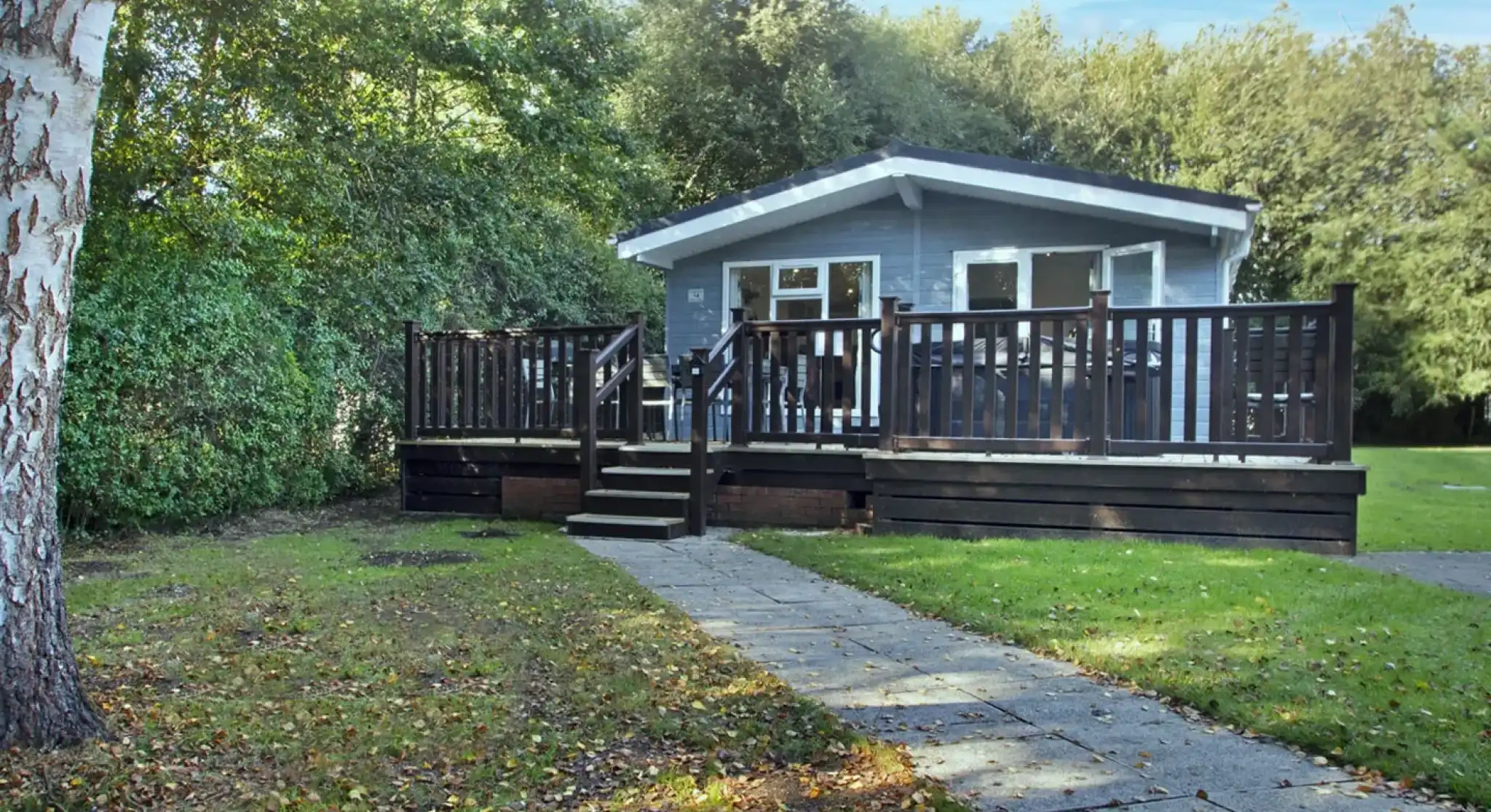 A cozy gray cabin with a dark wooden deck surrounded by greenery, featuring steps leading up to the entrance. A pathway made of stone tiles is visible in the foreground.