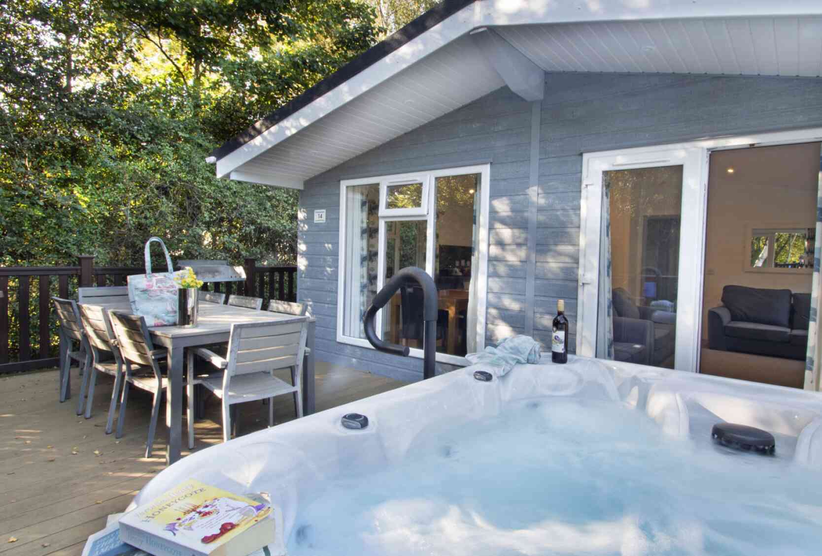 A hot tub with bubbling water is positioned on a wooden deck next to a dining table and chairs. Behind it, a cozy gray cabin features large windows and a door leading inside, surrounded by trees. A bottle of wine and a book are nearby on the deck.