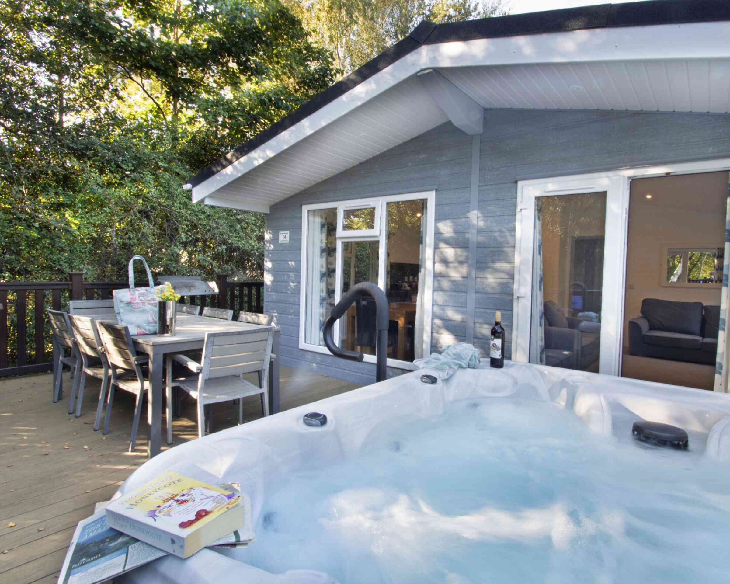 A hot tub with bubbling water is positioned on a wooden deck next to a dining table and chairs. Behind it, a cozy gray cabin features large windows and a door leading inside, surrounded by trees. A bottle of wine and a book are nearby on the deck.