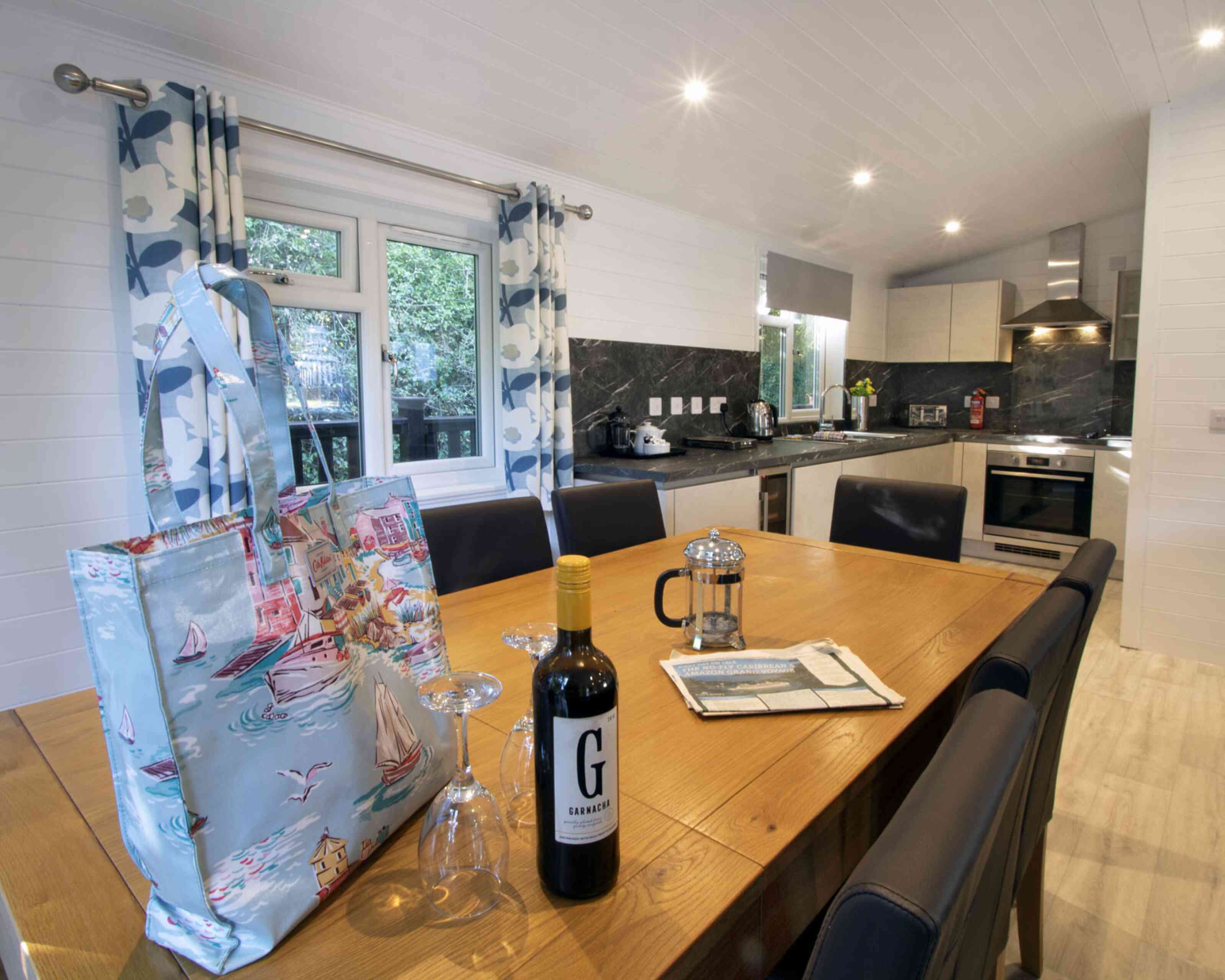 A modern kitchen and dining area featuring a wooden table set for a meal, a bottle of red wine, a French press, and glasses. Stylish curtains frame a window, while the kitchen showcases sleek cabinets and appliances against a dark marble backsplash. An attractive tote bag rests on the table.