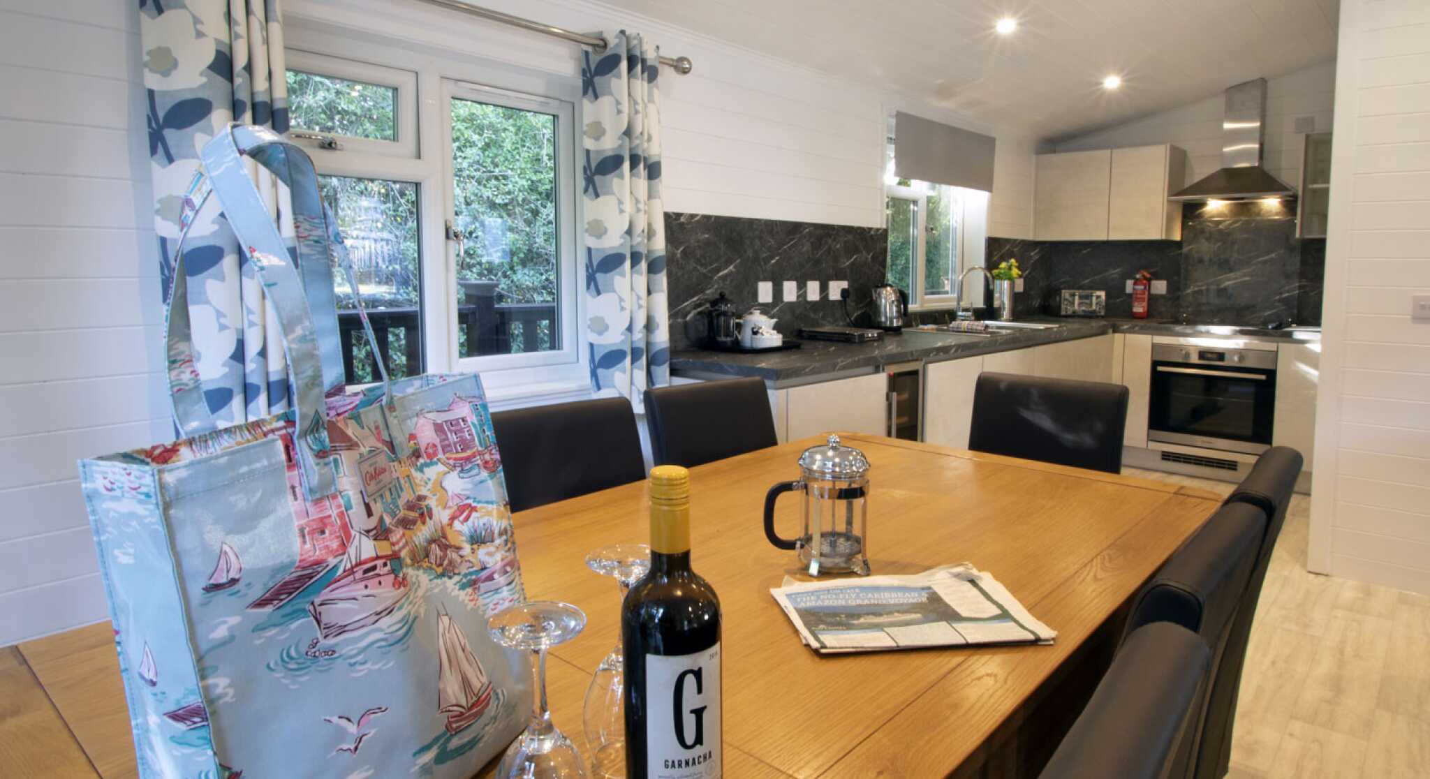 A modern kitchen and dining area featuring a wooden table set for a meal, a bottle of red wine, a French press, and glasses. Stylish curtains frame a window, while the kitchen showcases sleek cabinets and appliances against a dark marble backsplash. An attractive tote bag rests on the table.