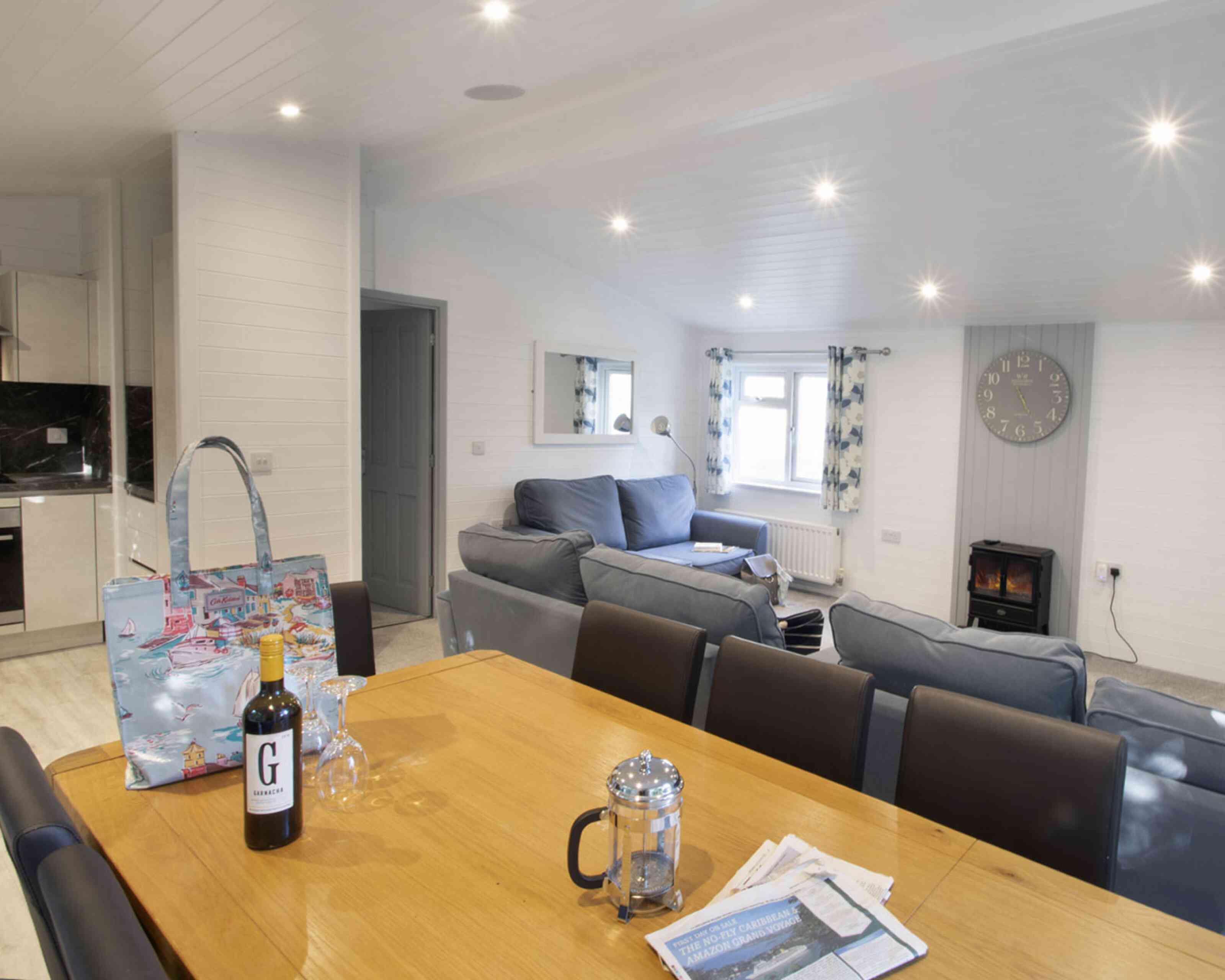 A cozy living space featuring a wooden dining table with wine and glassware, a comfortable grey sofa, a stylish handbag on the table, and a wall clock. A fireplace is visible in the background, along with curtains and large windows that allow natural light to brighten the room.