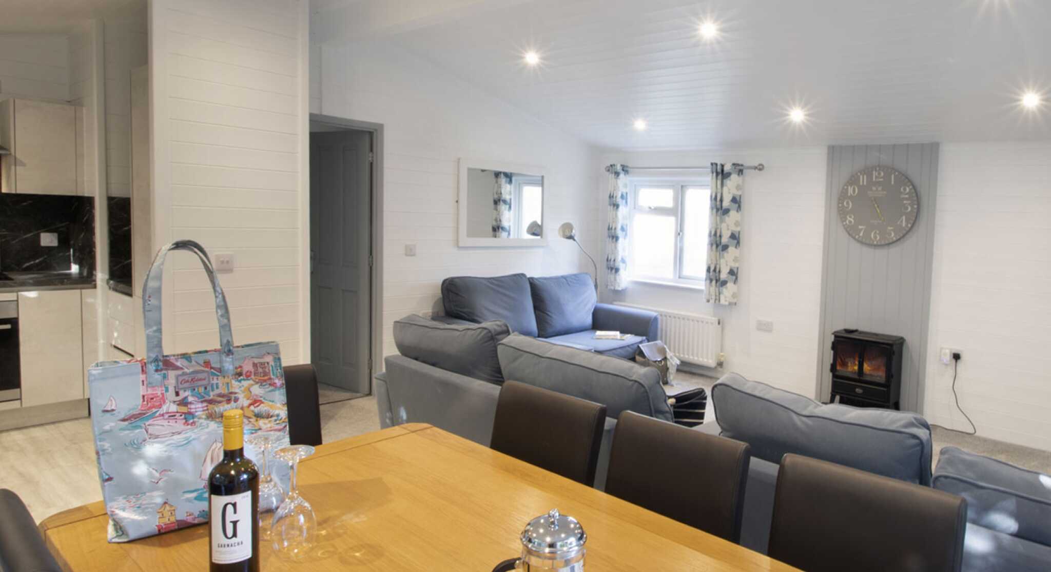 A cozy living space featuring a wooden dining table with wine and glassware, a comfortable grey sofa, a stylish handbag on the table, and a wall clock. A fireplace is visible in the background, along with curtains and large windows that allow natural light to brighten the room.