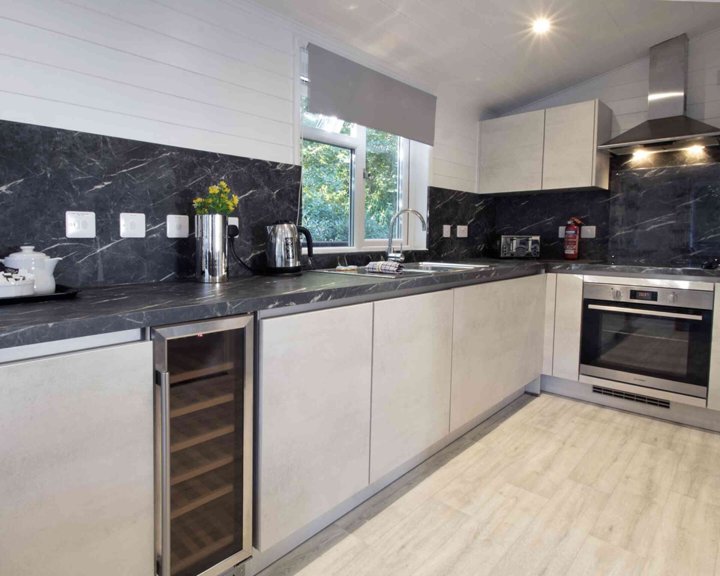Modern kitchen featuring sleek gray cabinets, a stainless steel oven, and a wine refrigerator. Natural light filters in through a window, highlighting the dark marble countertops and minimalist decor.
