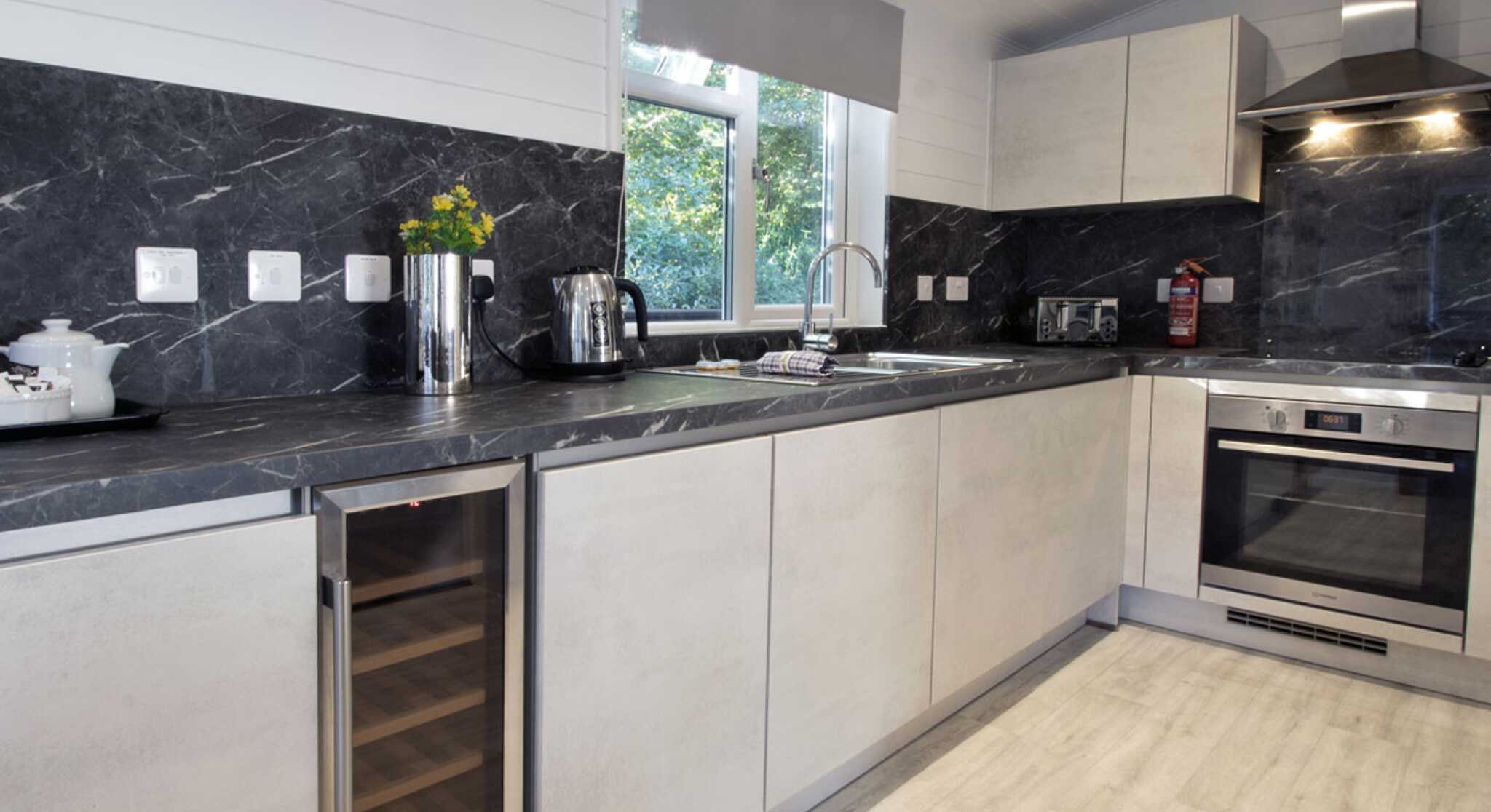 Modern kitchen featuring sleek gray cabinets, a stainless steel oven, and a wine refrigerator. Natural light filters in through a window, highlighting the dark marble countertops and minimalist decor.