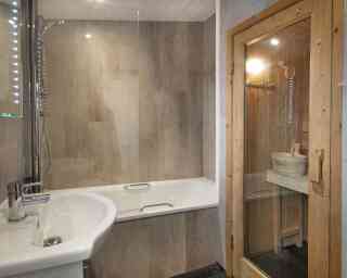 A modern bathroom featuring a glass shower enclosure and a bathtub. A wooden sauna is visible in the corner, with a wooden bucket and ladle inside. The walls have a sleek, stone-like finish and soft lighting illuminates the space.