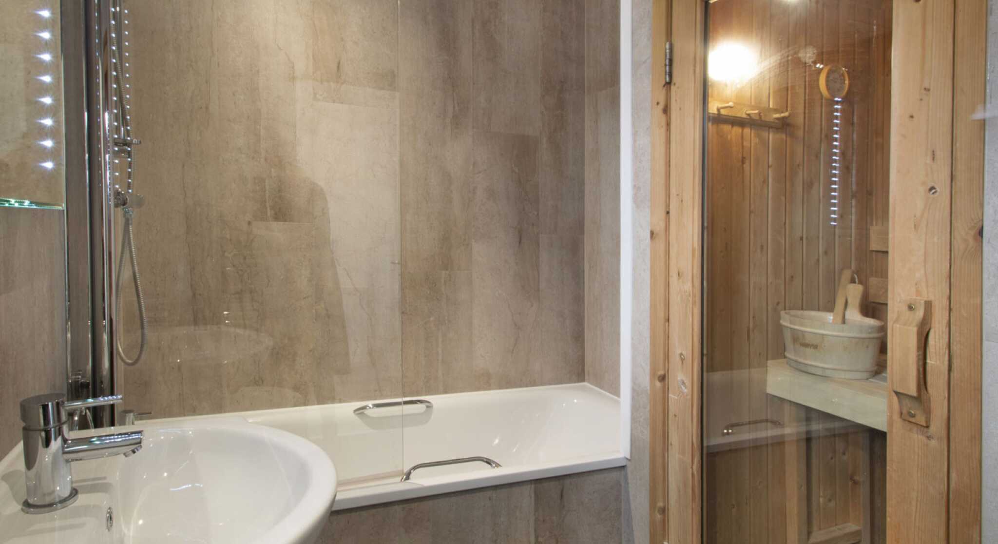 A modern bathroom featuring a glass shower enclosure and a bathtub. A wooden sauna is visible in the corner, with a wooden bucket and ladle inside. The walls have a sleek, stone-like finish and soft lighting illuminates the space.