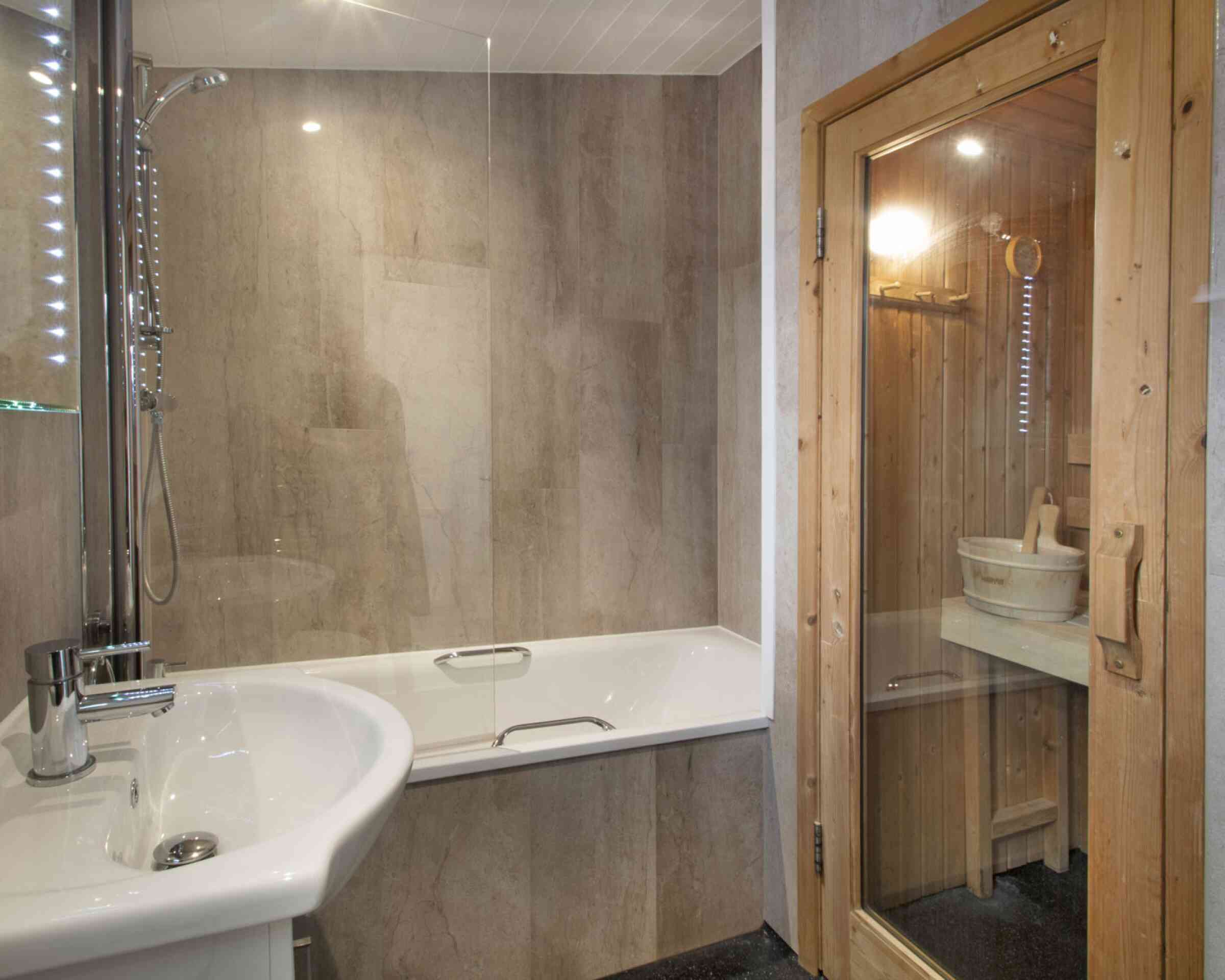 A modern bathroom featuring a glass shower enclosure and a bathtub. A wooden sauna is visible in the corner, with a wooden bucket and ladle inside. The walls have a sleek, stone-like finish and soft lighting illuminates the space.