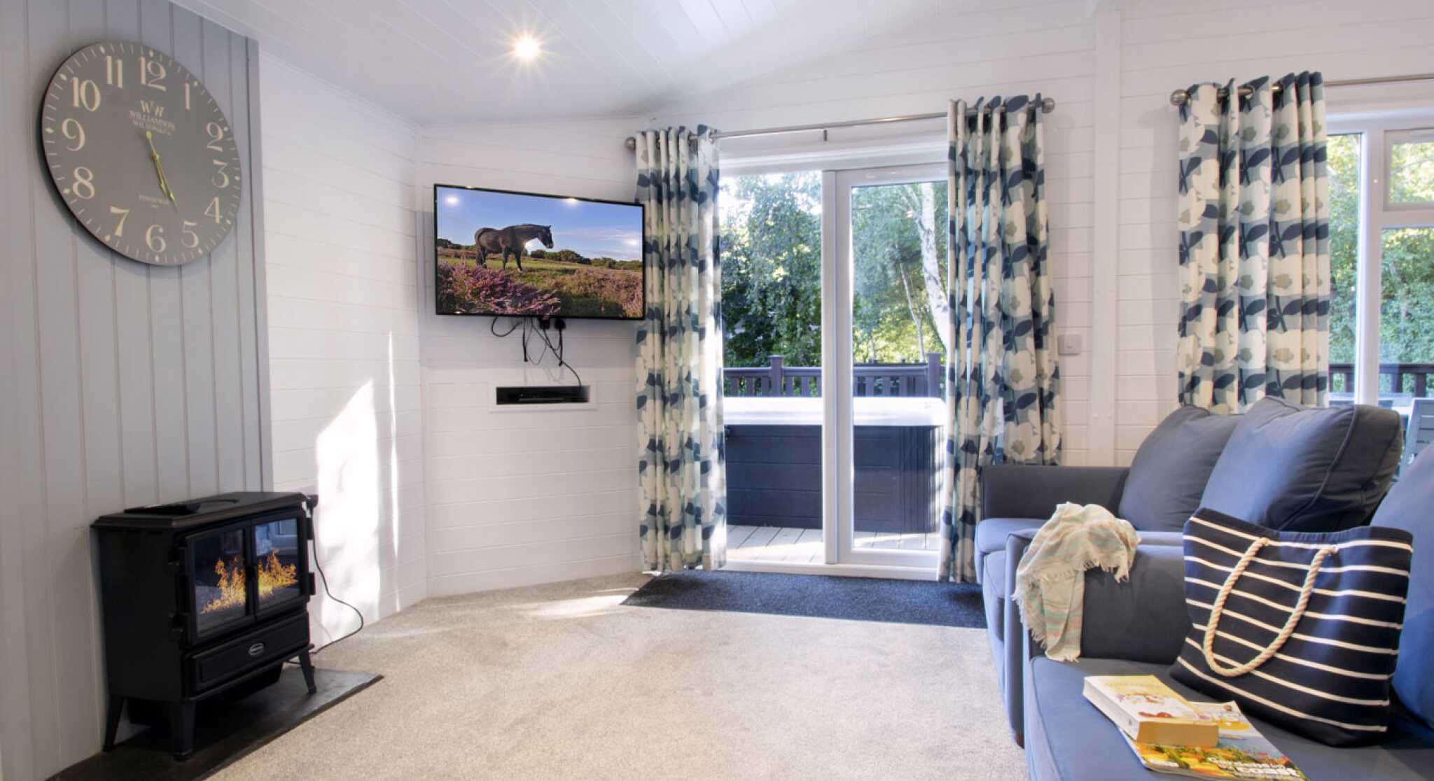 A cozy living room featuring a blue sofa with decorative pillows, a modern clock on the wall, and a flat-screen TV mounted high. Large windows with patterned curtains allow natural light to fill the space, while a black wood stove adds warmth. A small table with a magazine rests on the carpeted floor.