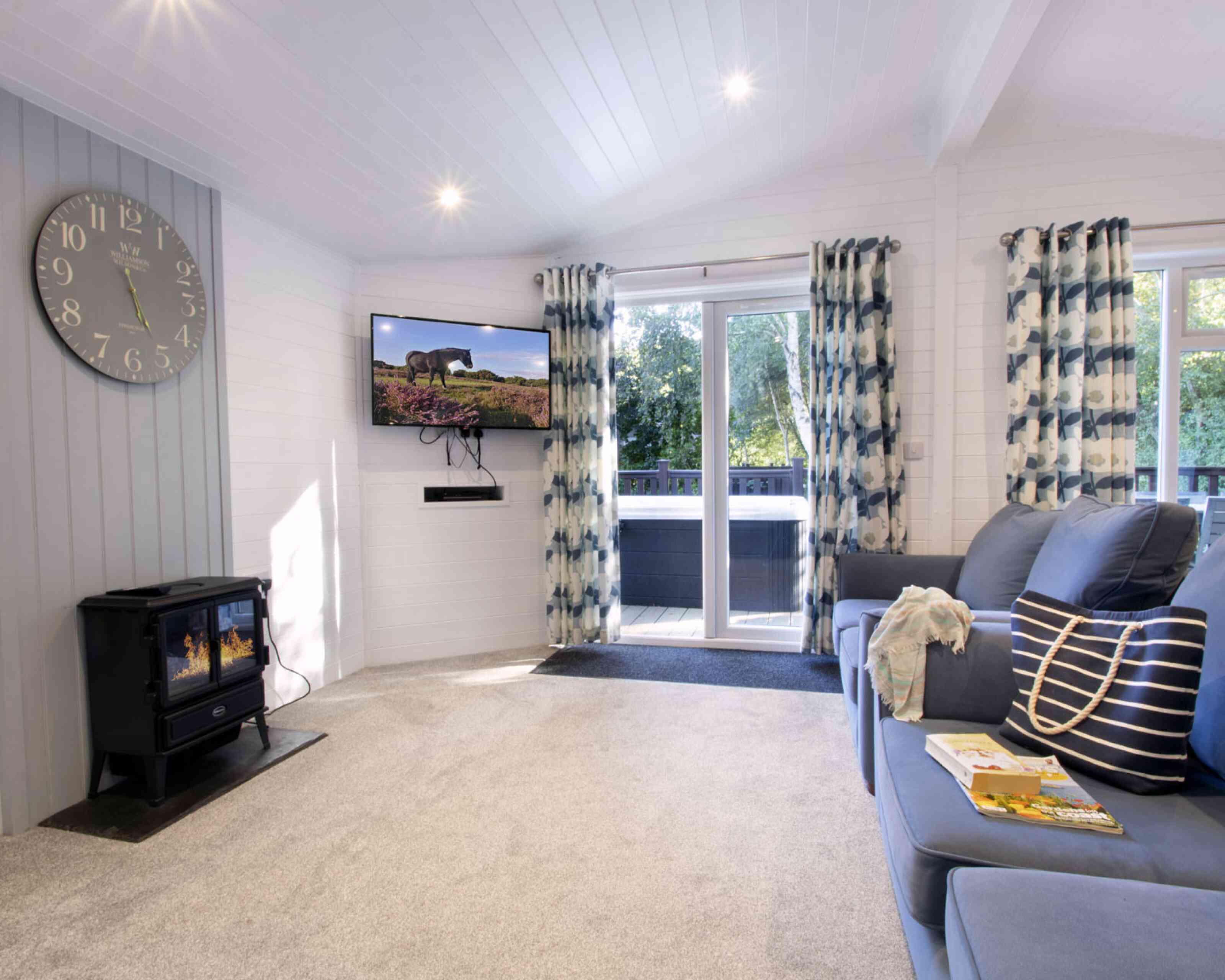 A cozy living room featuring a blue sofa with decorative pillows, a modern clock on the wall, and a flat-screen TV mounted high. Large windows with patterned curtains allow natural light to fill the space, while a black wood stove adds warmth. A small table with a magazine rests on the carpeted floor.