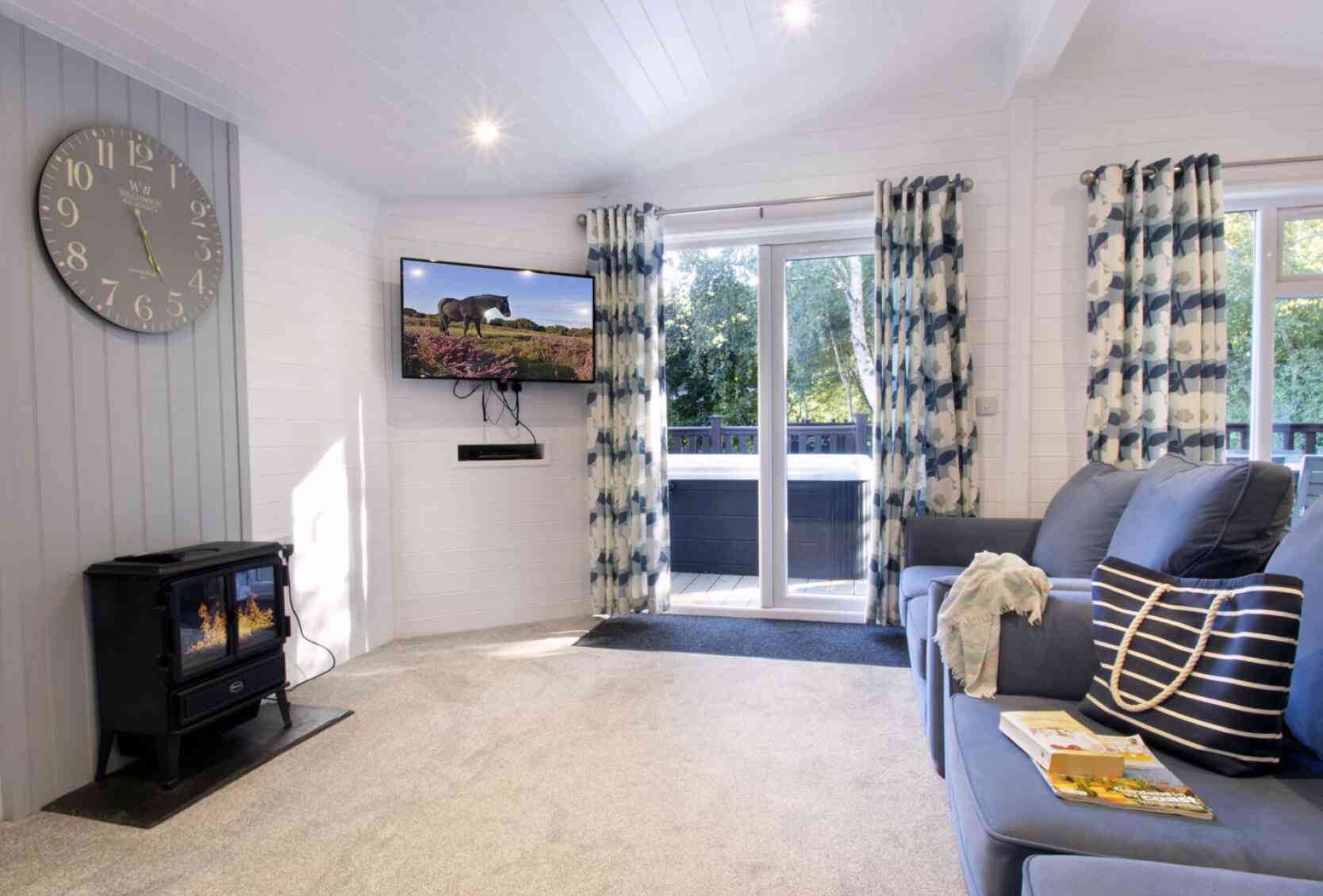 A cozy living room featuring a blue sofa with decorative pillows, a modern clock on the wall, and a flat-screen TV mounted high. Large windows with patterned curtains allow natural light to fill the space, while a black wood stove adds warmth. A small table with a magazine rests on the carpeted floor.