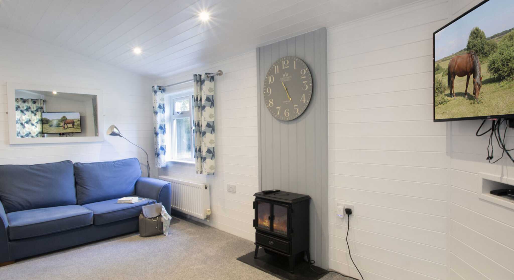 Cozy living room featuring a blue sofa, a wall clock, a television displaying a horse, and a fireplace on the floor. The room has light-colored walls and curtains with floral patterns.