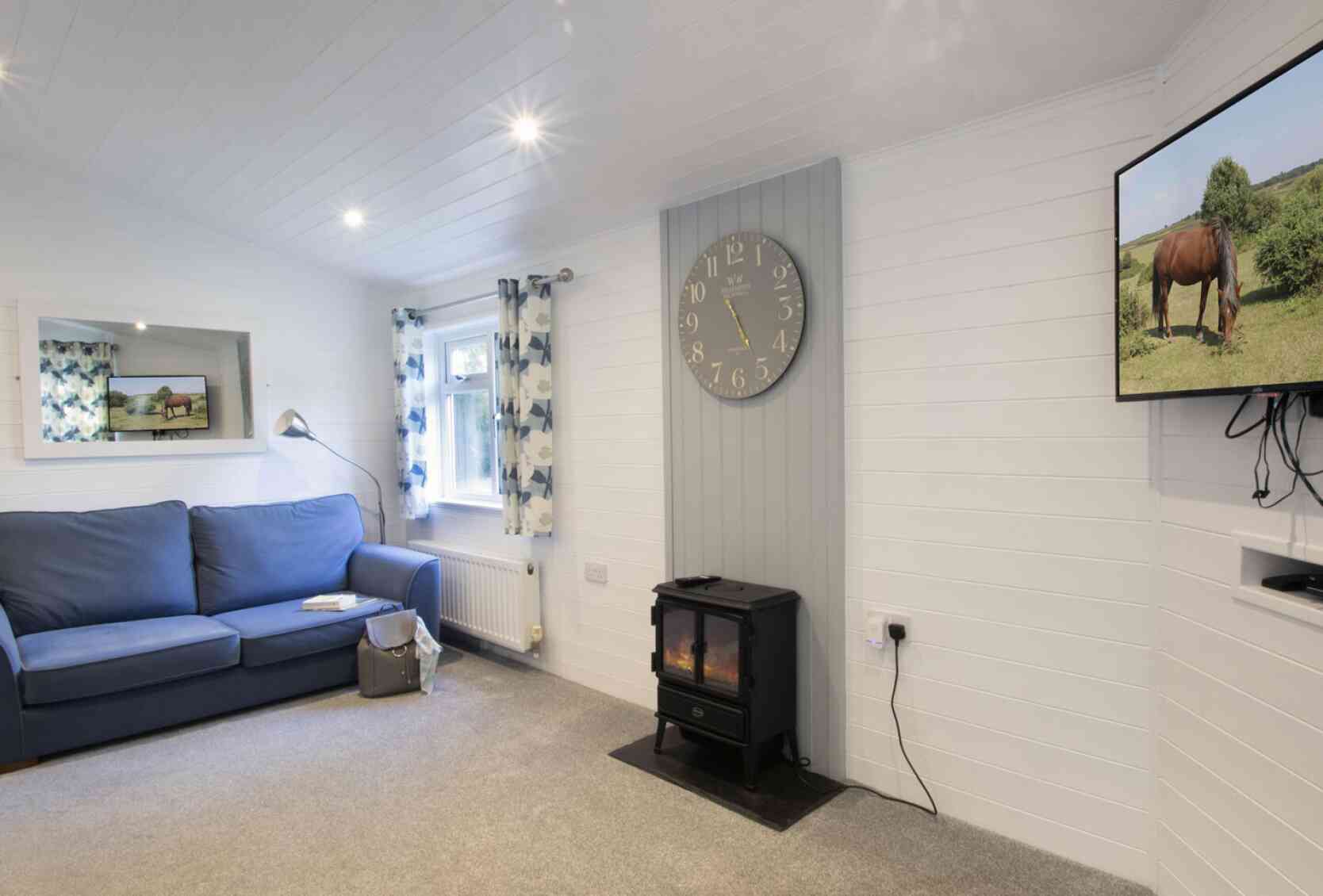 Cozy living room featuring a blue sofa, a wall clock, a television displaying a horse, and a fireplace on the floor. The room has light-colored walls and curtains with floral patterns.