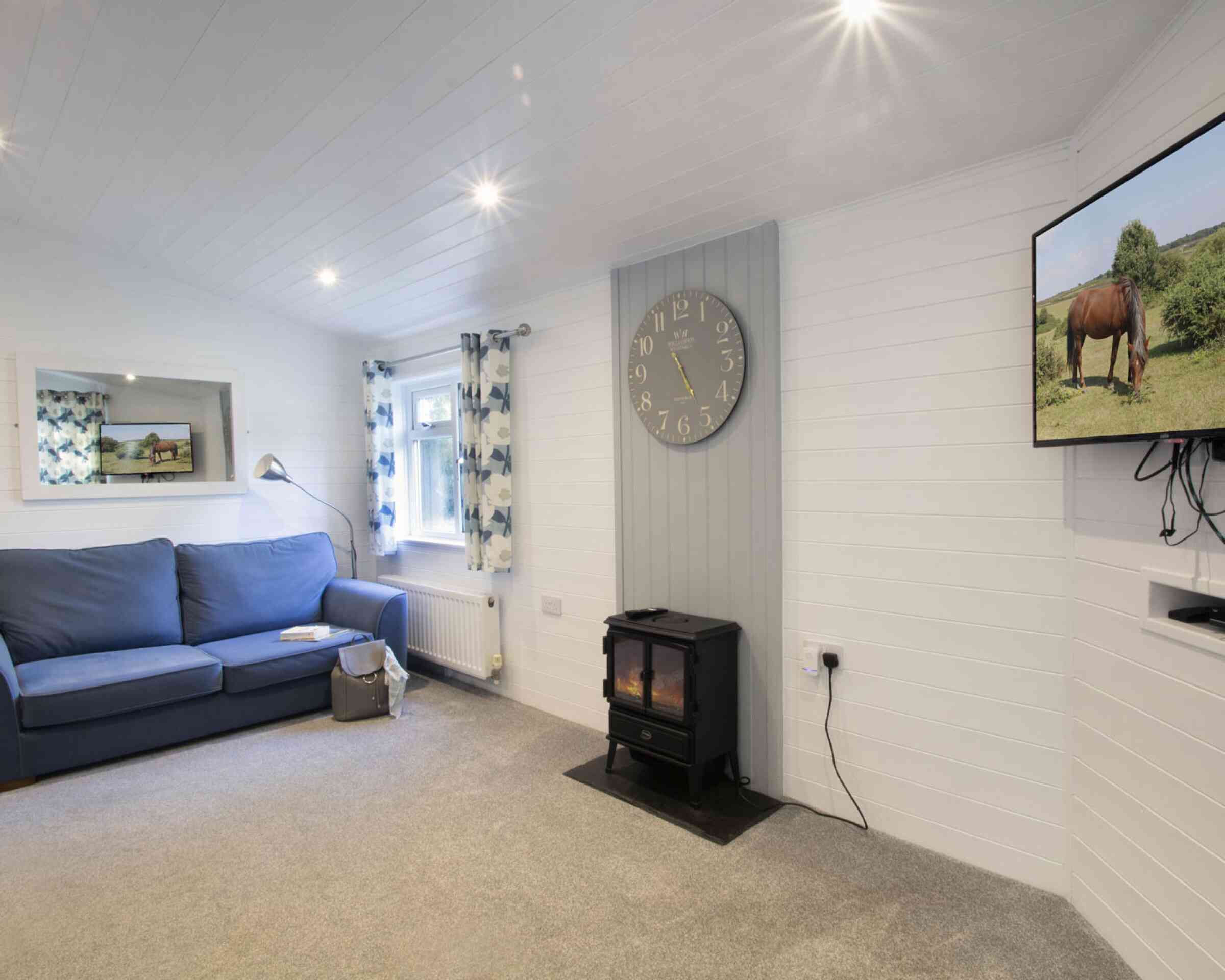 Cozy living room featuring a blue sofa, a wall clock, a television displaying a horse, and a fireplace on the floor. The room has light-colored walls and curtains with floral patterns.