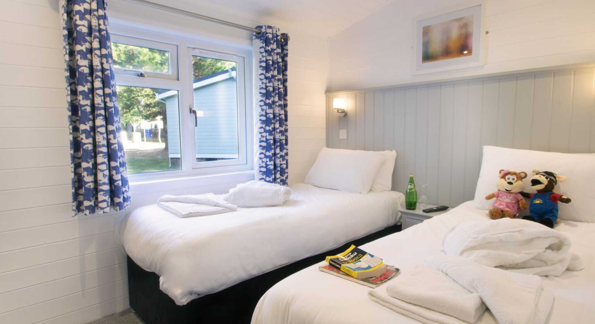 A cozy bedroom featuring two single beds with white bedding. A window with decorative blue curtains allows natural light in. Each bed has neatly placed towels, and stuffed animals are arranged on one of the beds. A small stack of magazines rests on the other bed, and the walls are painted in light colors, creating a welcoming atmosphere.