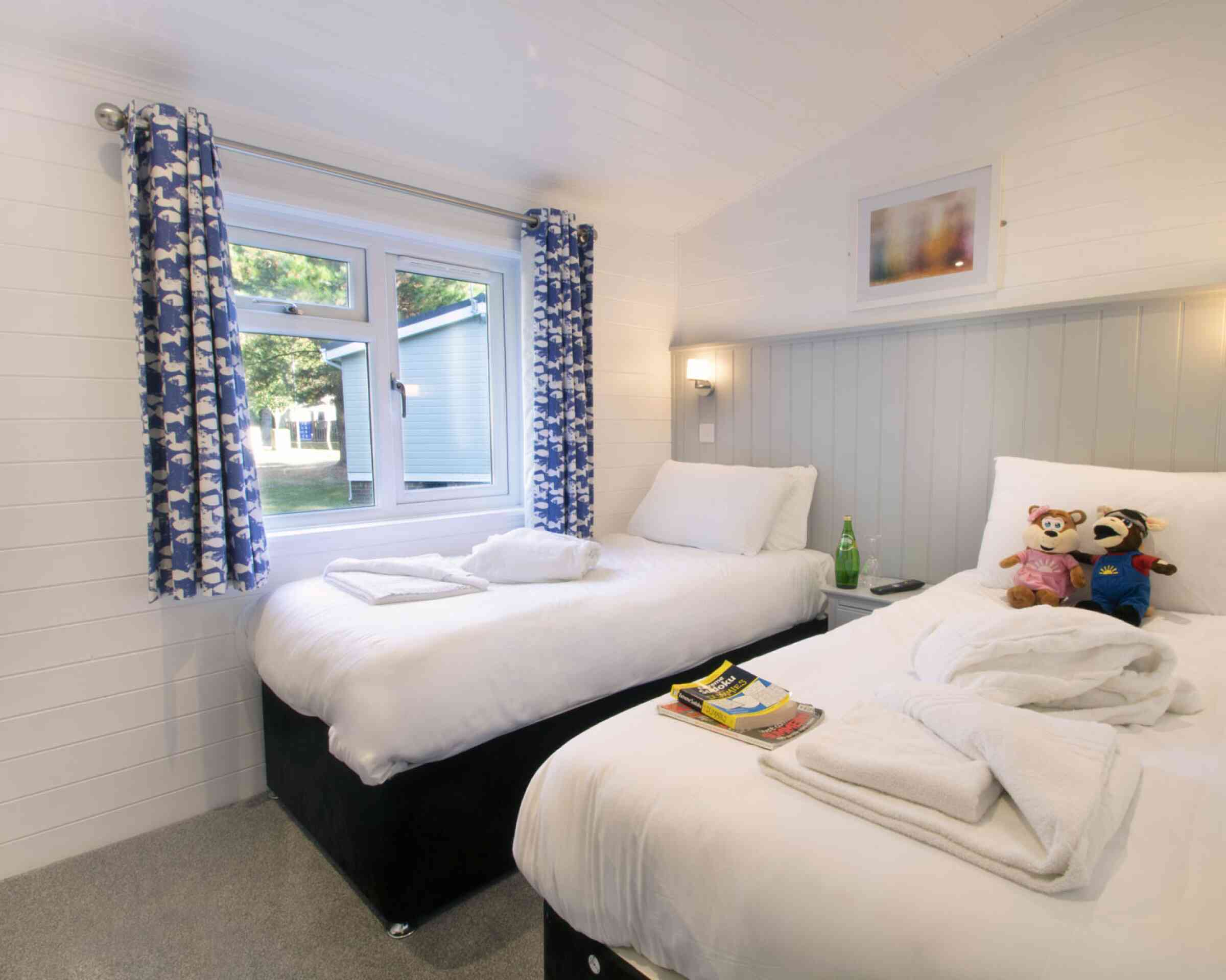 A cozy bedroom featuring two single beds with white bedding. A window with decorative blue curtains allows natural light in. Each bed has neatly placed towels, and stuffed animals are arranged on one of the beds. A small stack of magazines rests on the other bed, and the walls are painted in light colors, creating a welcoming atmosphere.