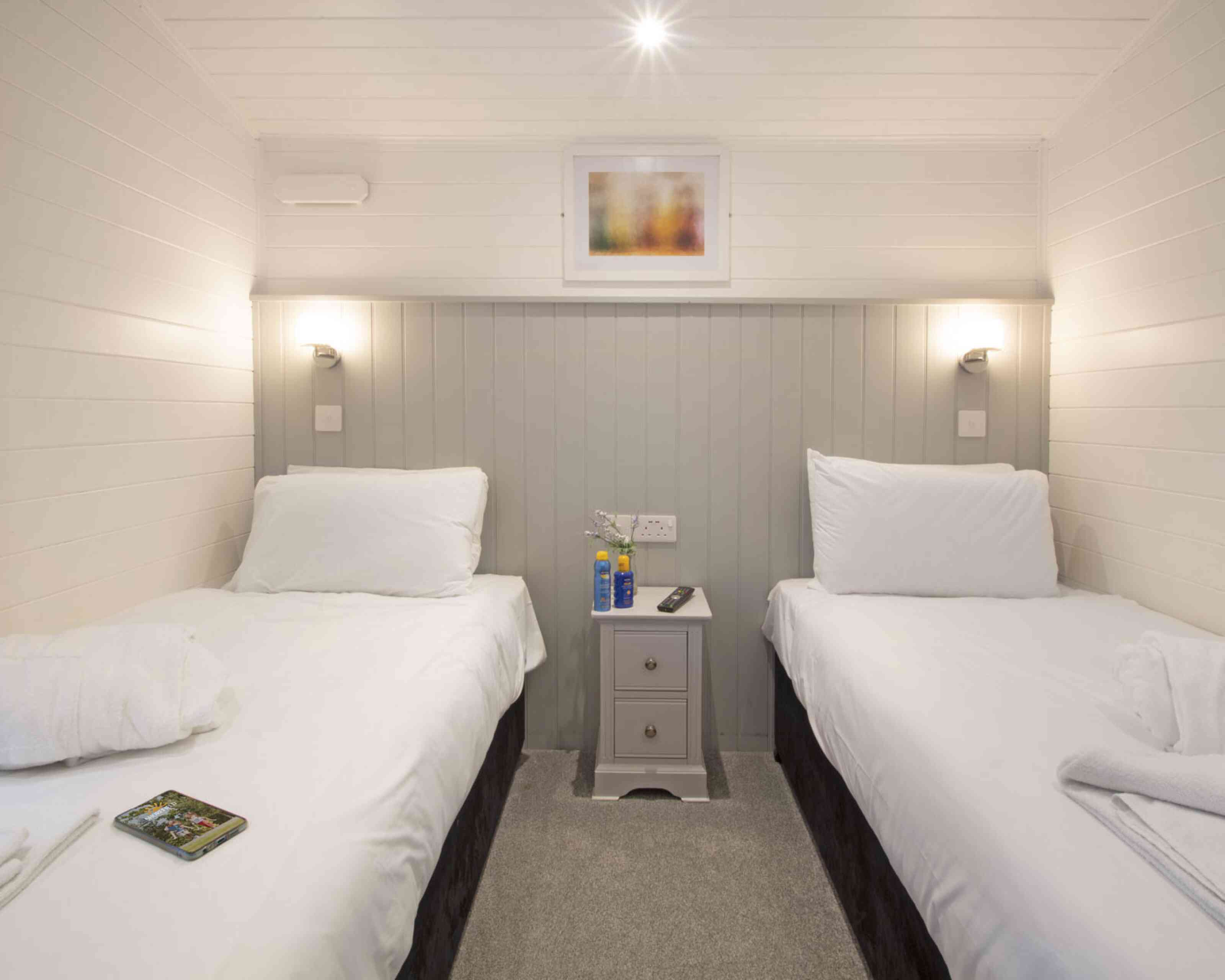 A cozy bedroom featuring two single beds with white linens, a small nightstand in between, and modern wall sconces. The walls are painted in light colors with a framed abstract artwork above the nightstand. A soft gray carpet completes the inviting atmosphere.