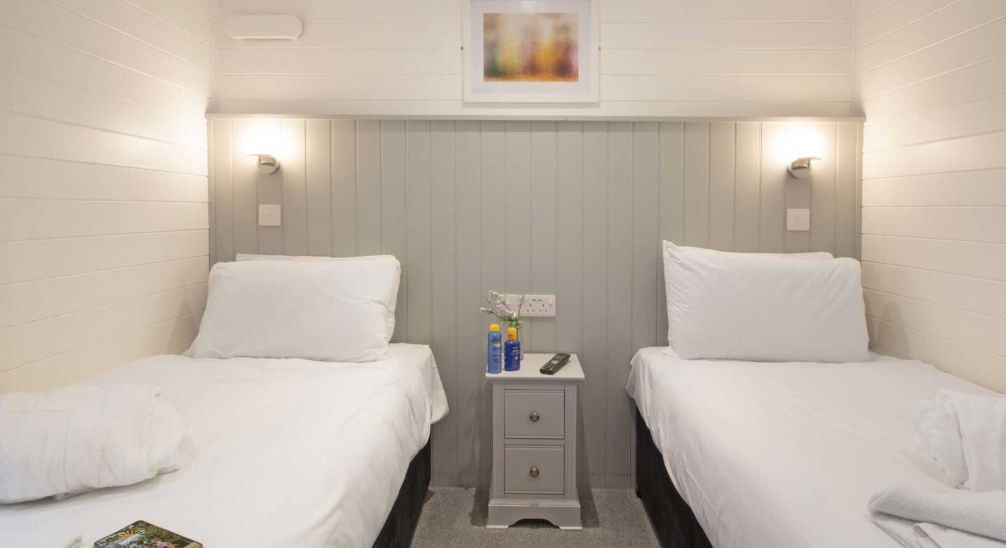 A cozy bedroom featuring two single beds with white linens, a small nightstand in between, and modern wall sconces. The walls are painted in light colors with a framed abstract artwork above the nightstand. A soft gray carpet completes the inviting atmosphere.