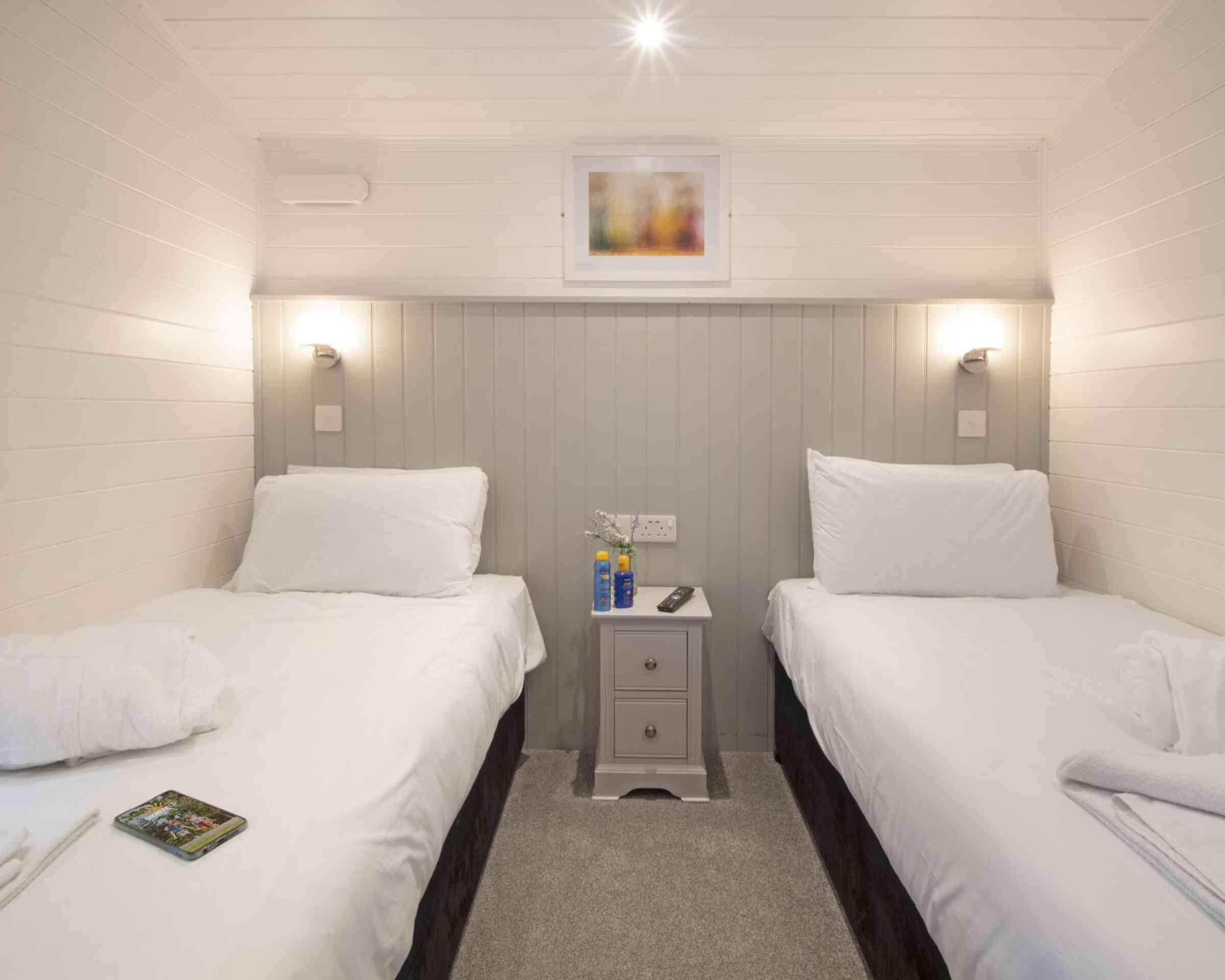 A cozy bedroom featuring two single beds with white linens, a small nightstand in between, and modern wall sconces. The walls are painted in light colors with a framed abstract artwork above the nightstand. A soft gray carpet completes the inviting atmosphere.