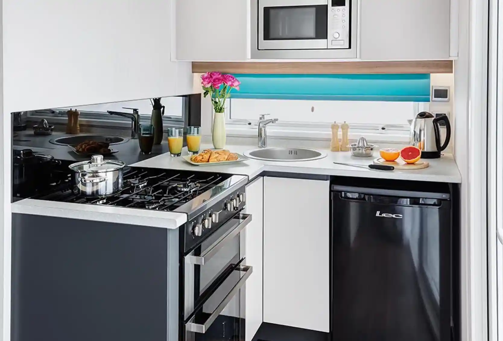 A modern kitchen featuring a black refrigerator, a stainless steel microwave above a gas stove, and a circular sink. The countertop is minimalistic, with a vase of flowers and breakfast items like croissants and oranges arranged nearby. Bright blue accents complete the contemporary design.