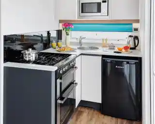 A modern kitchen featuring a black refrigerator, a stainless steel microwave above a gas stove, and a circular sink. The countertop is minimalistic, with a vase of flowers and breakfast items like croissants and oranges arranged nearby. Bright blue accents complete the contemporary design.