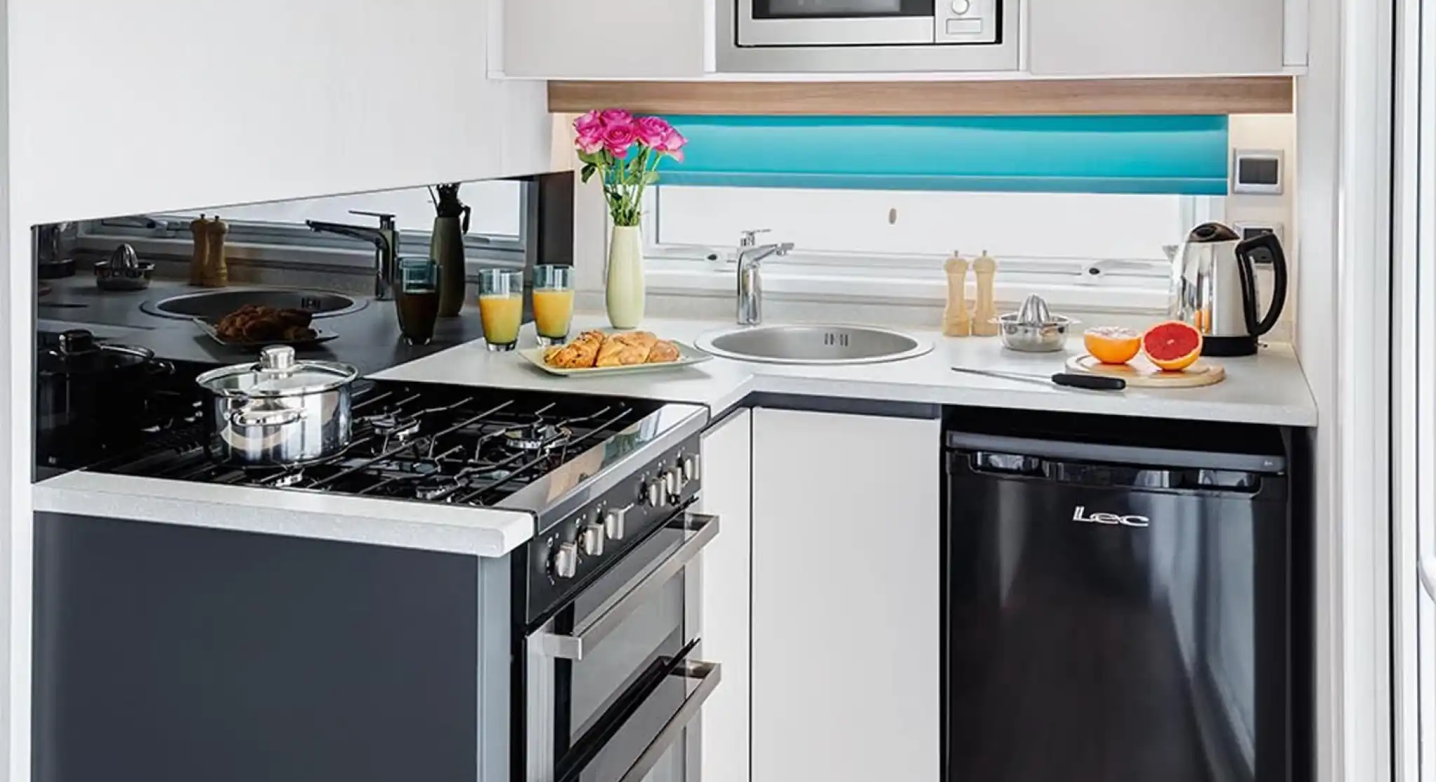 A modern kitchen featuring a black refrigerator, a stainless steel microwave above a gas stove, and a circular sink. The countertop is minimalistic, with a vase of flowers and breakfast items like croissants and oranges arranged nearby. Bright blue accents complete the contemporary design.