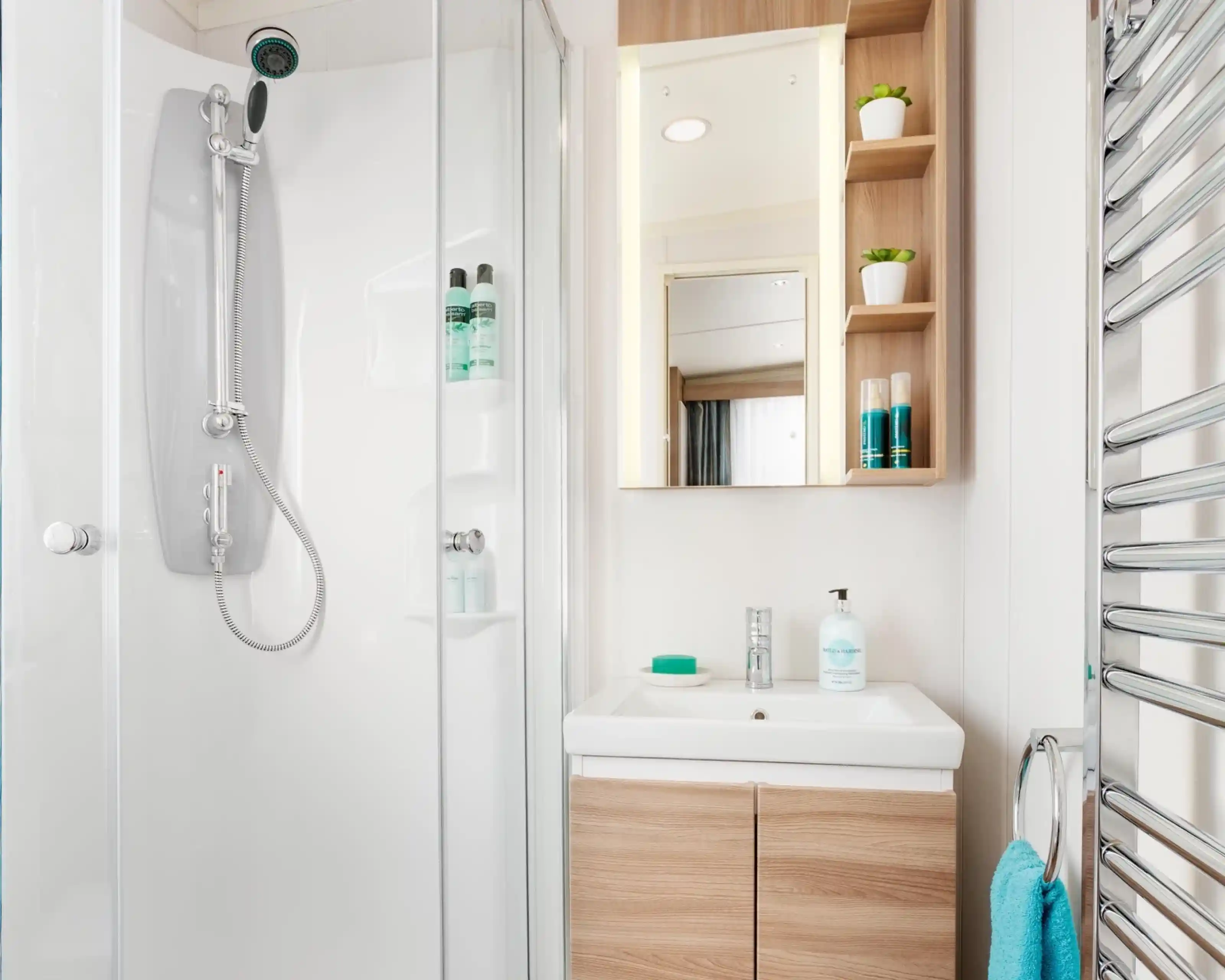 A modern bathroom featuring a glass shower enclosure with a rainfall showerhead. A sleek vanity with a white sink and wooden cabinets is visible, along with a large mirror above it. Shelves with green plants and toiletries are mounted on the wall. A blue towel hangs on a heated towel rail.