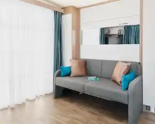 A modern living room features a gray sofa with turquoise and bronze pillows, set against a backdrop of sheer white curtains and light wooden flooring. A mirror and a small TV are visible in the reflection. A magazine lies on the sofa.