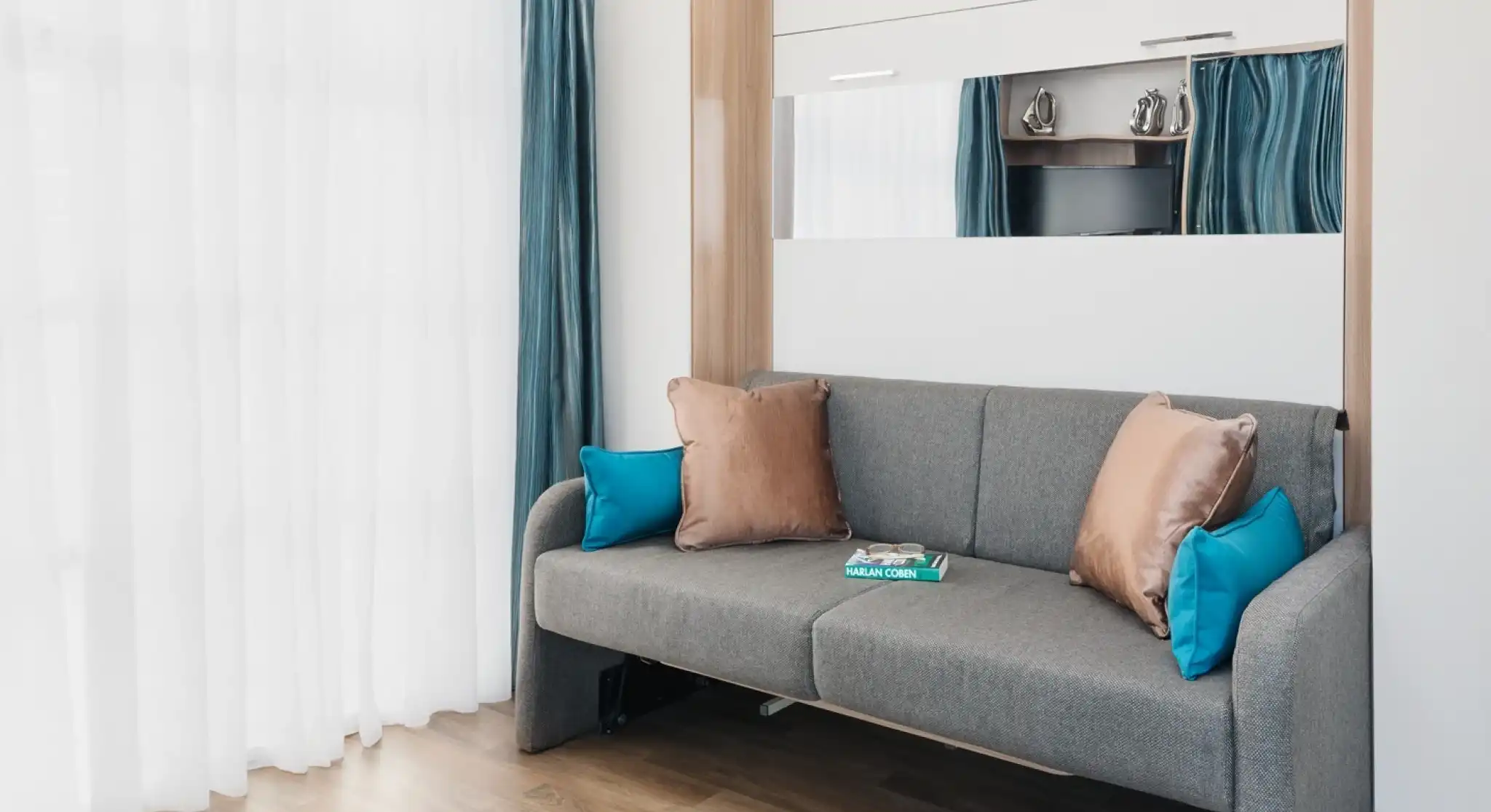 A modern living room features a gray sofa with turquoise and bronze pillows, set against a backdrop of sheer white curtains and light wooden flooring. A mirror and a small TV are visible in the reflection. A magazine lies on the sofa.