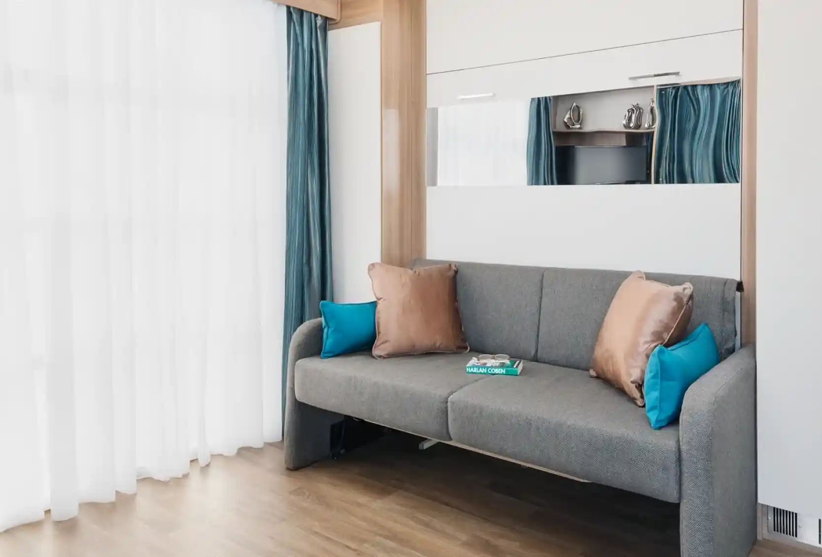 A modern living room features a gray sofa with turquoise and bronze pillows, set against a backdrop of sheer white curtains and light wooden flooring. A mirror and a small TV are visible in the reflection. A magazine lies on the sofa.