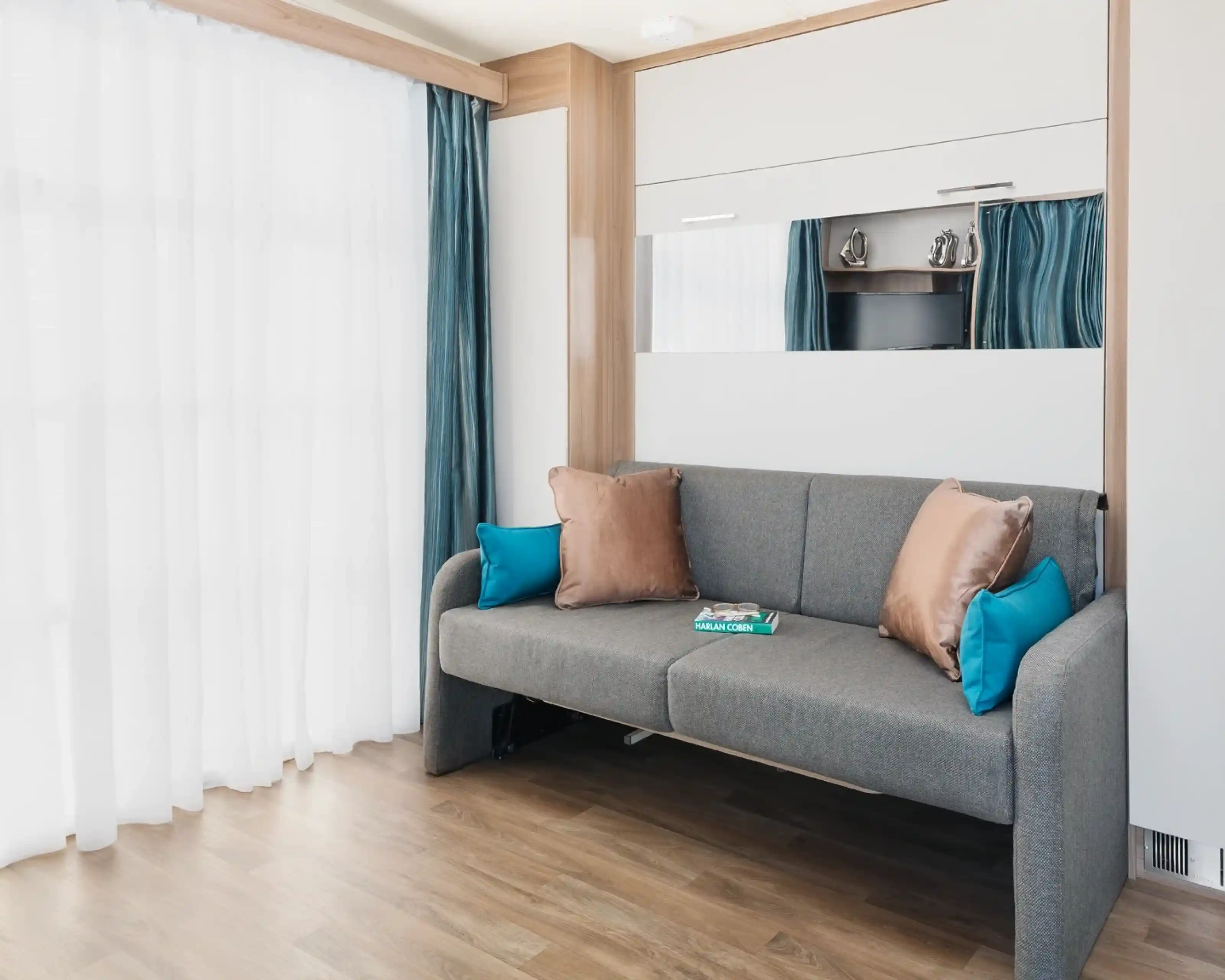 A modern living room features a gray sofa with turquoise and bronze pillows, set against a backdrop of sheer white curtains and light wooden flooring. A mirror and a small TV are visible in the reflection. A magazine lies on the sofa.