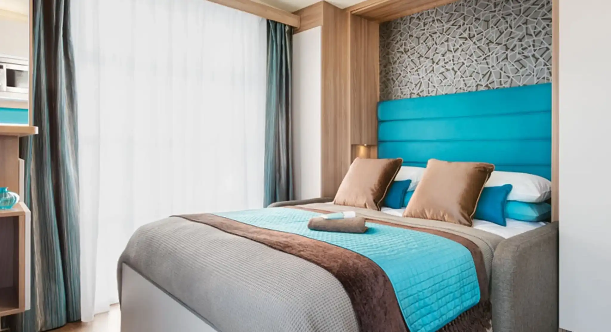 Modern bedroom featuring a queen-sized bed with blue and brown accents. A patterned feature wall complements the light wood cabinetry. Large windows with sheer curtains allow natural light to filter in. Decorative pillows and a throw blanket add a cozy touch.