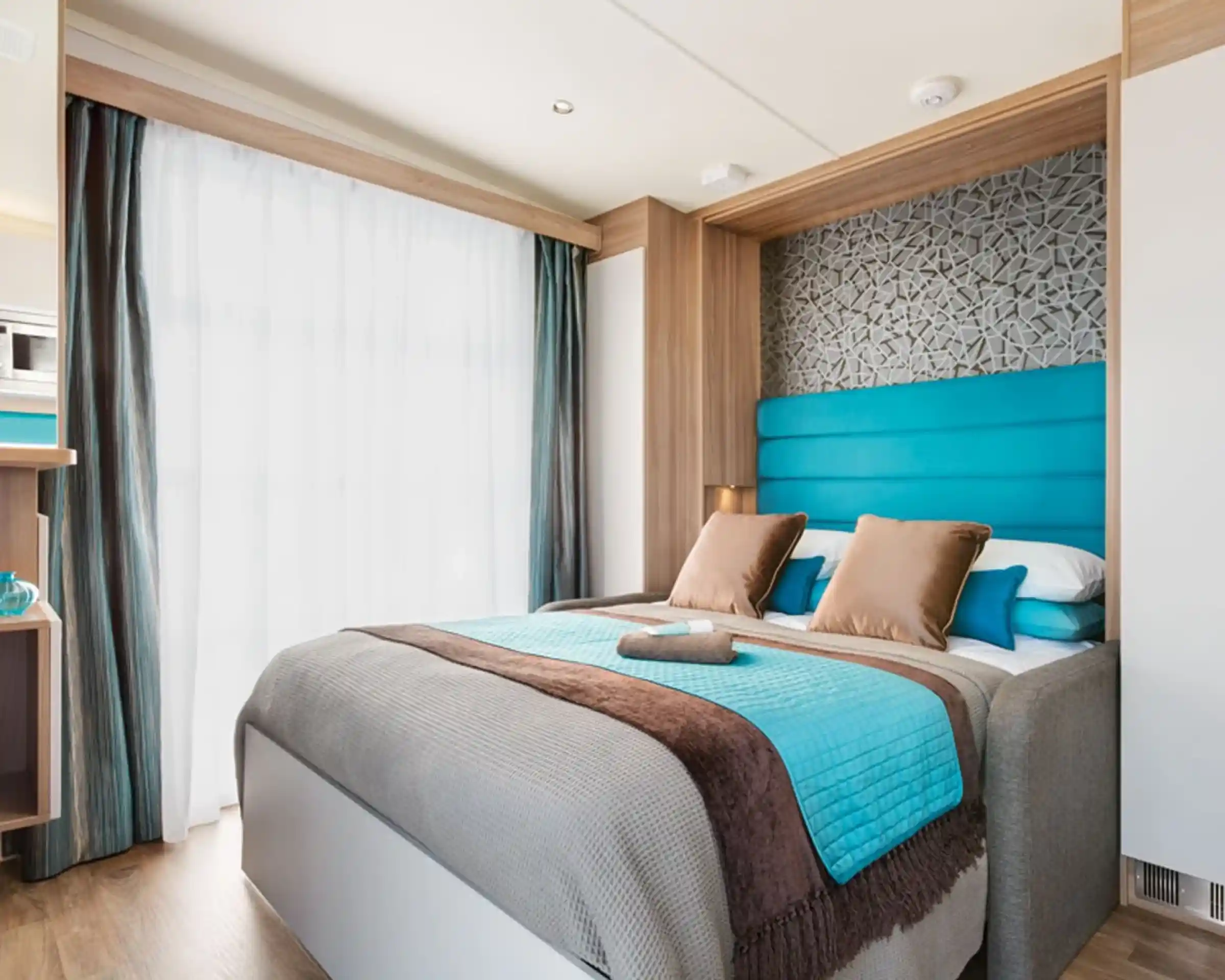 Modern bedroom featuring a queen-sized bed with blue and brown accents. A patterned feature wall complements the light wood cabinetry. Large windows with sheer curtains allow natural light to filter in. Decorative pillows and a throw blanket add a cozy touch.