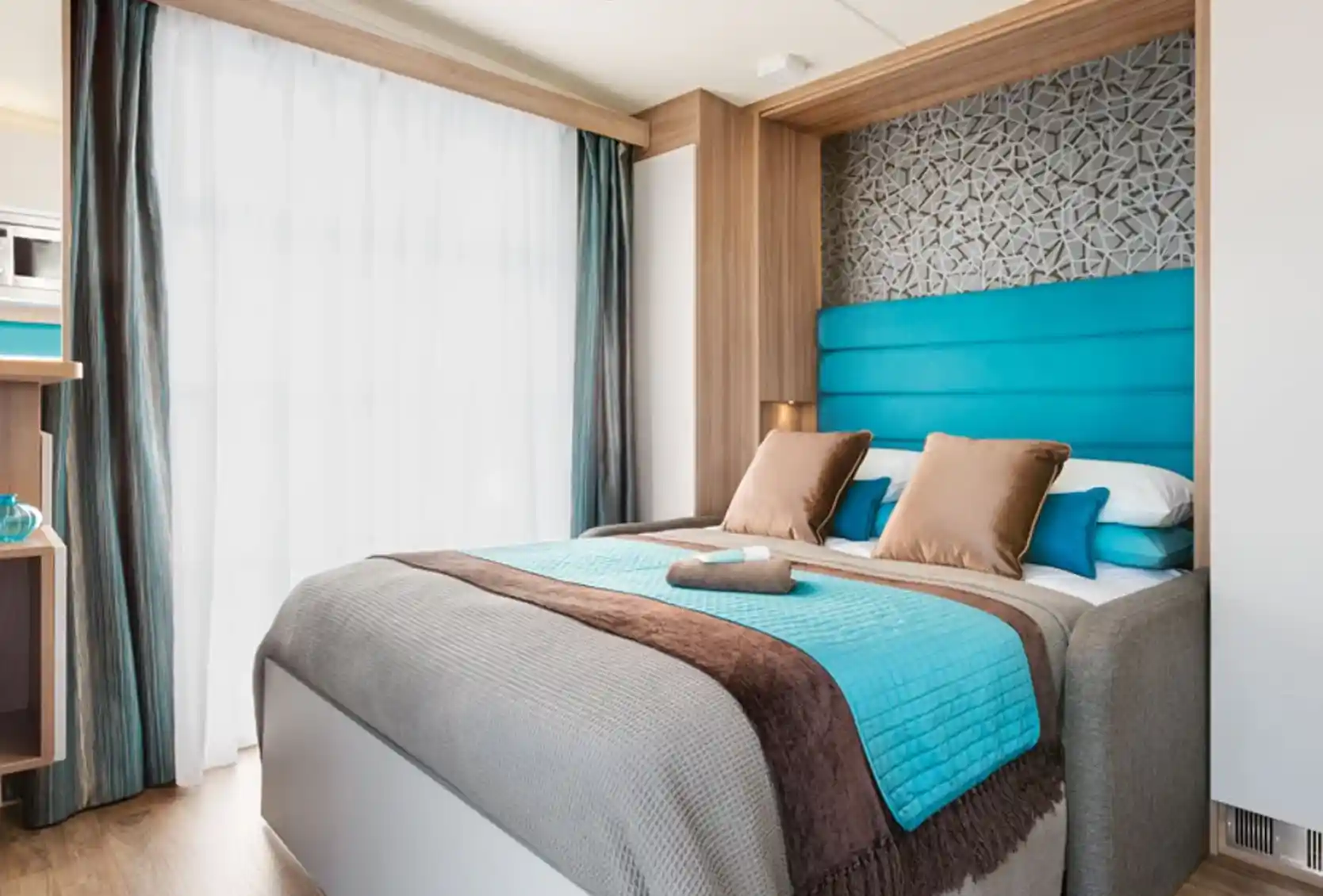 Modern bedroom featuring a queen-sized bed with blue and brown accents. A patterned feature wall complements the light wood cabinetry. Large windows with sheer curtains allow natural light to filter in. Decorative pillows and a throw blanket add a cozy touch.