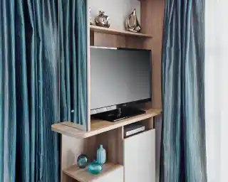 A sleek wooden entertainment unit featuring a flat-screen television, blue decorative vases, and stylish ornaments. The unit is flanked by luxurious, flowing blue curtains.
