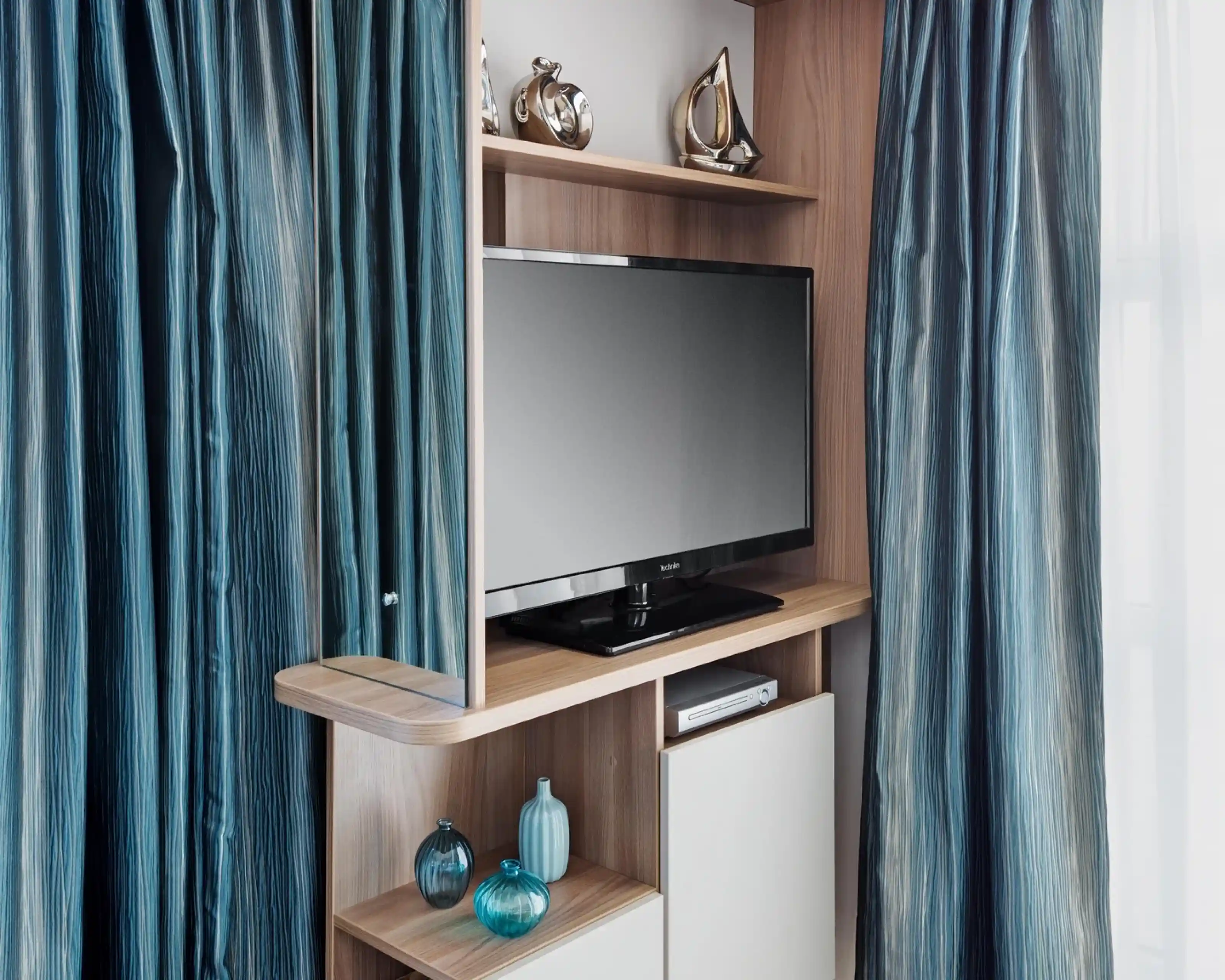 A sleek wooden entertainment unit featuring a flat-screen television, blue decorative vases, and stylish ornaments. The unit is flanked by luxurious, flowing blue curtains.