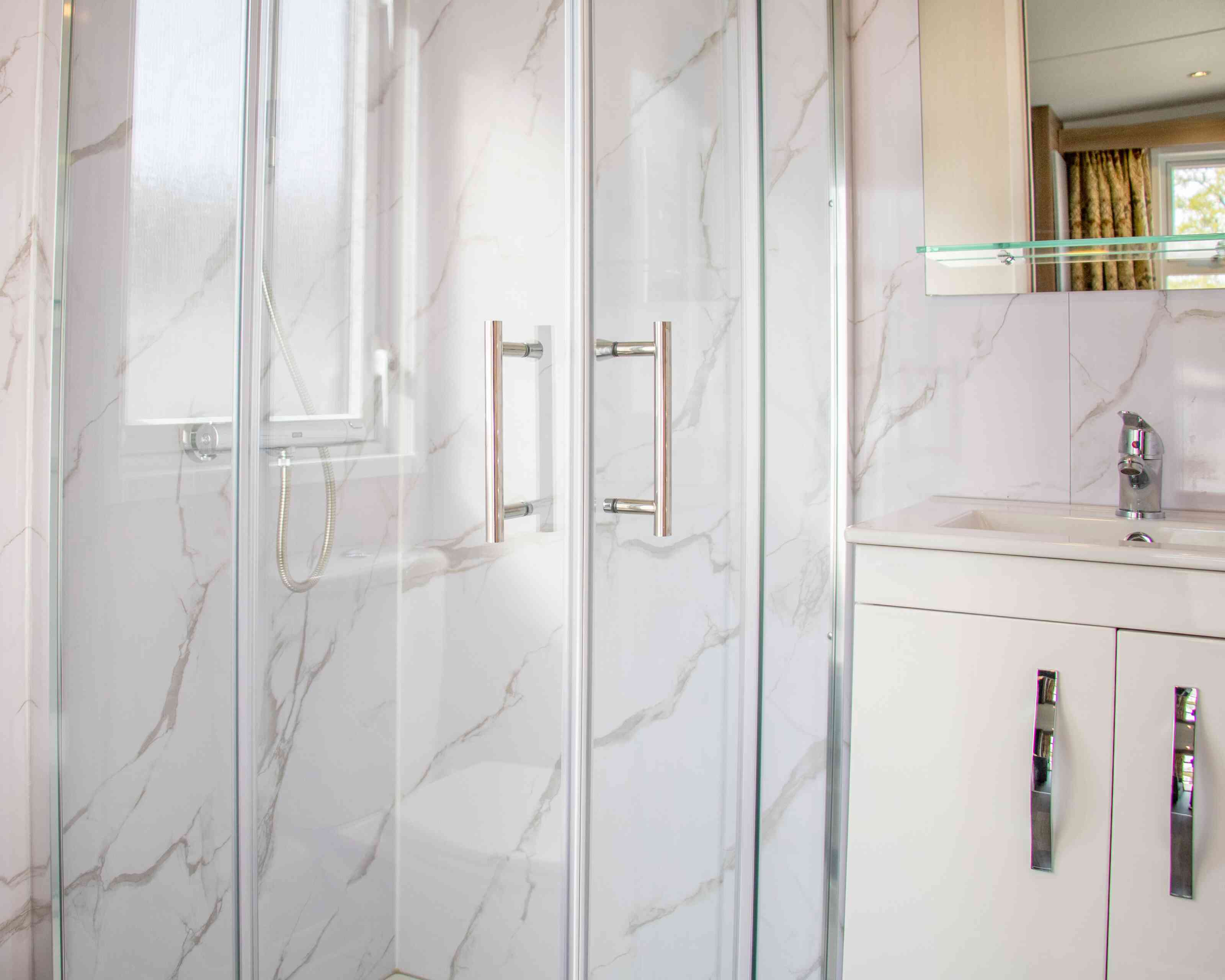 A modern bathroom featuring a glass shower enclosure with a marble-patterned wall, a sleek white vanity with silver handles, and a polished faucet. Natural light enters through a nearby window.
