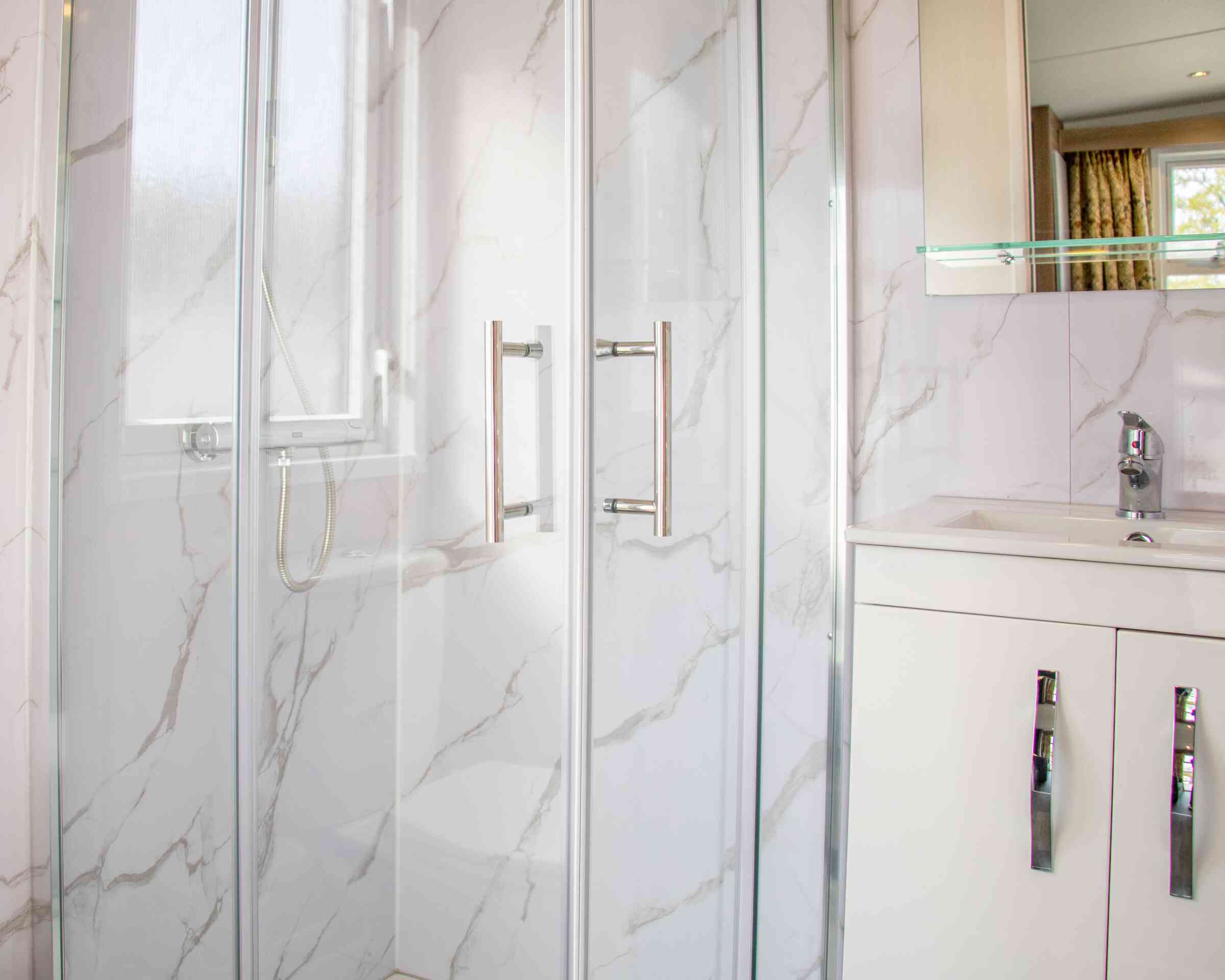 A modern bathroom featuring a glass shower enclosure with a marble-patterned wall, a sleek white vanity with silver handles, and a polished faucet. Natural light enters through a nearby window.