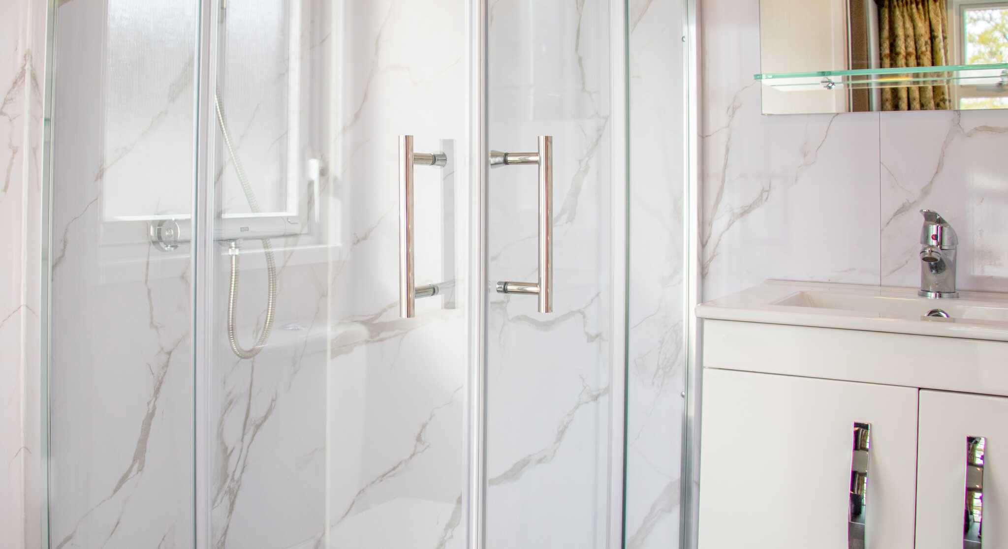 A modern bathroom featuring a glass shower enclosure with a marble-patterned wall, a sleek white vanity with silver handles, and a polished faucet. Natural light enters through a nearby window.