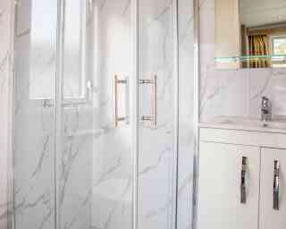 A modern bathroom featuring a glass shower enclosure with a marble-patterned wall, a sleek white vanity with silver handles, and a polished faucet. Natural light enters through a nearby window.