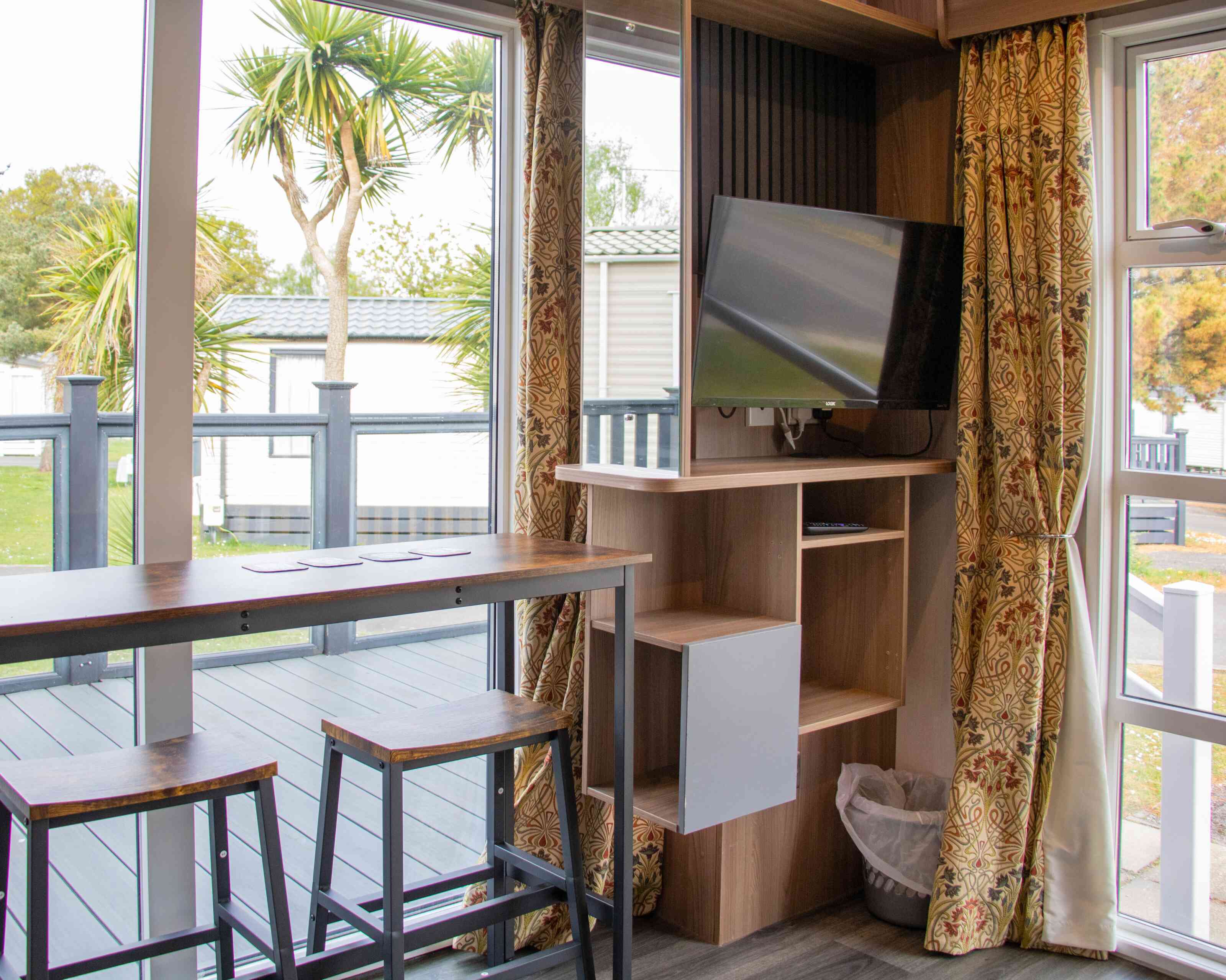 A cozy interior of a small living space featuring a wooden bar with three stools, a mounted television on a wall unit, and large windows with curtains. Outside, palm trees and a deck are visible.