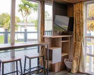 A cozy interior of a small living space featuring a wooden bar with three stools, a mounted television on a wall unit, and large windows with curtains. Outside, palm trees and a deck are visible.