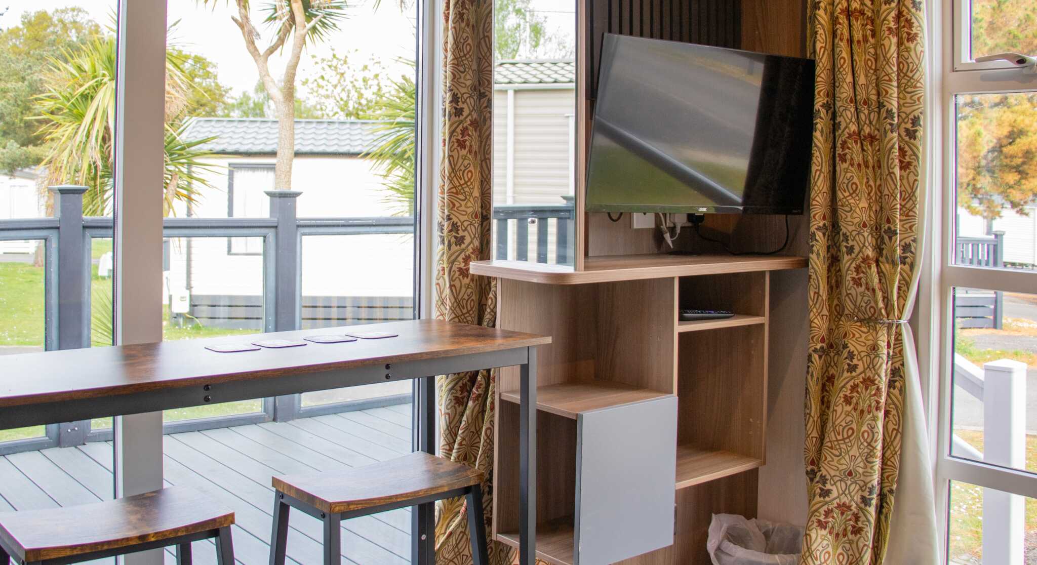 A cozy interior of a small living space featuring a wooden bar with three stools, a mounted television on a wall unit, and large windows with curtains. Outside, palm trees and a deck are visible.