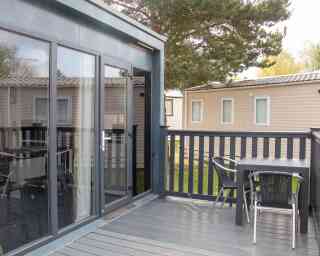 A modern deck area featuring a table and chairs, with large glass doors leading to an interior space. In the background, there are several beige cabins and green trees.